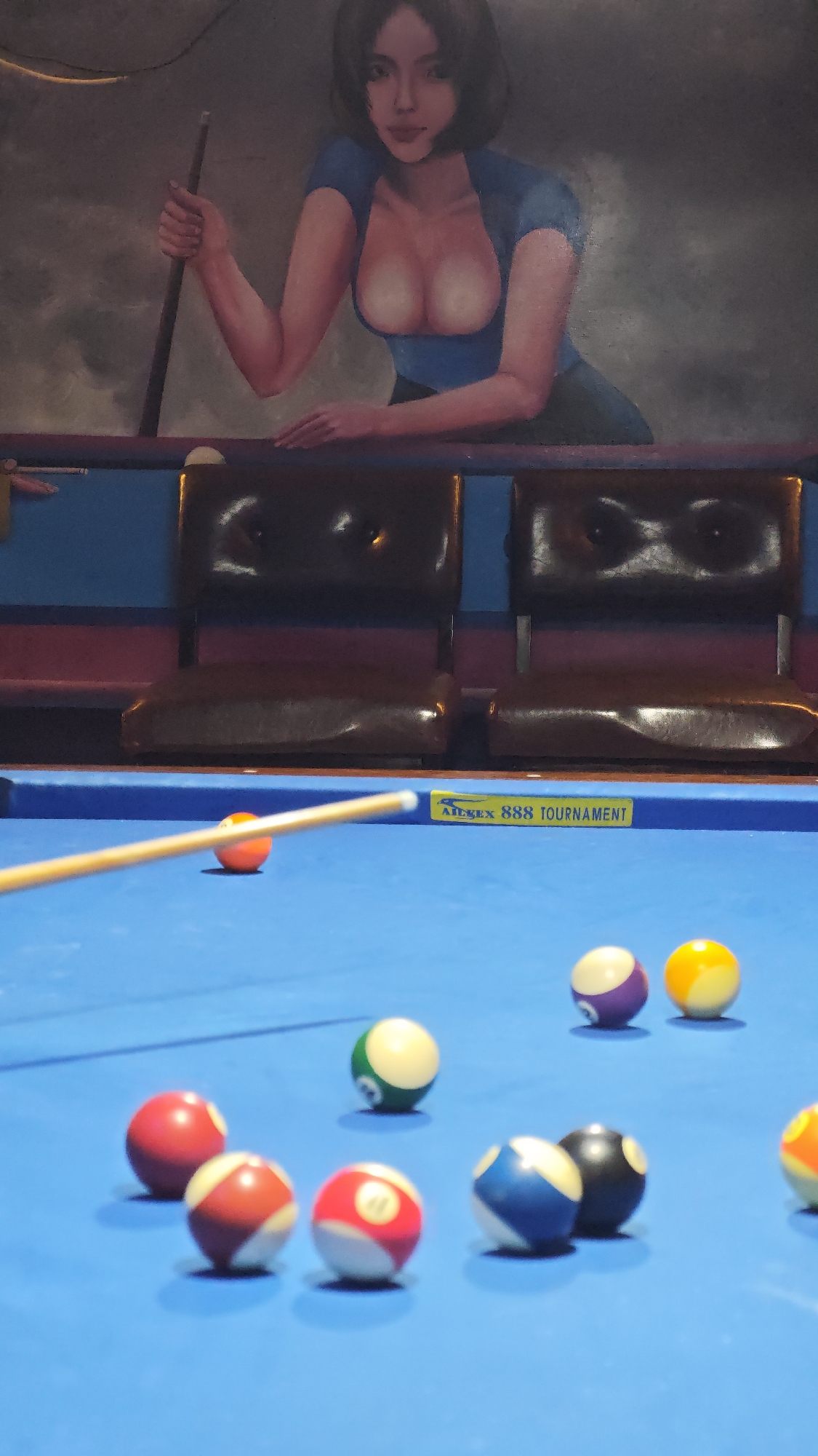 The image depicts a billiards table in a room with a painting of a woman above. The billiards table is set up with multiple pool balls in view, and a cue stick aiming towards one. The room appears to be indoors with chairs visible behind the billiards table. There is no evidence of the location being in Đà Nẵng, Vietnam.