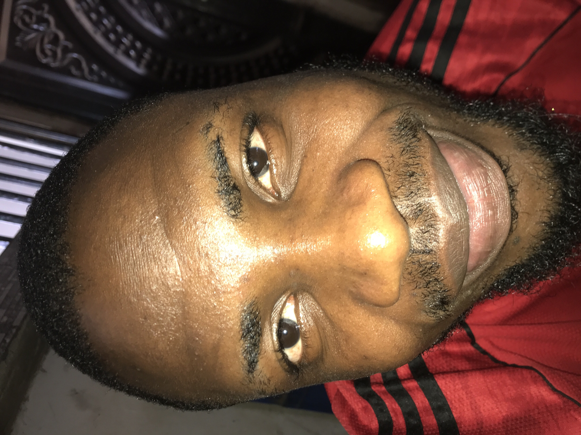 This media file features a close-up portrait of a person, likely male, with dark skin, short dark hair, and a dark beard and mustache. The image is oriented sideways, with the person's head tilted 90 degrees to the left. The individual is looking directly at the camera with an open-mouthed expression, revealing their tongue, which suggests a moment of laughter, excitement, or playful communication.
The person is wearing a red garment with distinctive black stripes, indicative of athletic or leisure wear, possibly a t-shirt or jersey. The skin on their face appears somewhat shiny, which could be due to perspiration or natural skin oils.
The background is dark and out of focus. Hints of architectural details are visible in the upper left, possibly a decorative ceiling or a dark structure. The lower left shows a faint, lighter, striped pattern. The overall lighting suggests an indoor setting or low-light conditions. There is no visible text in the image. Given the provided location context of "New Stadium, Nigeria," the attire and appearance might suggest the person is at or around a sporting event.