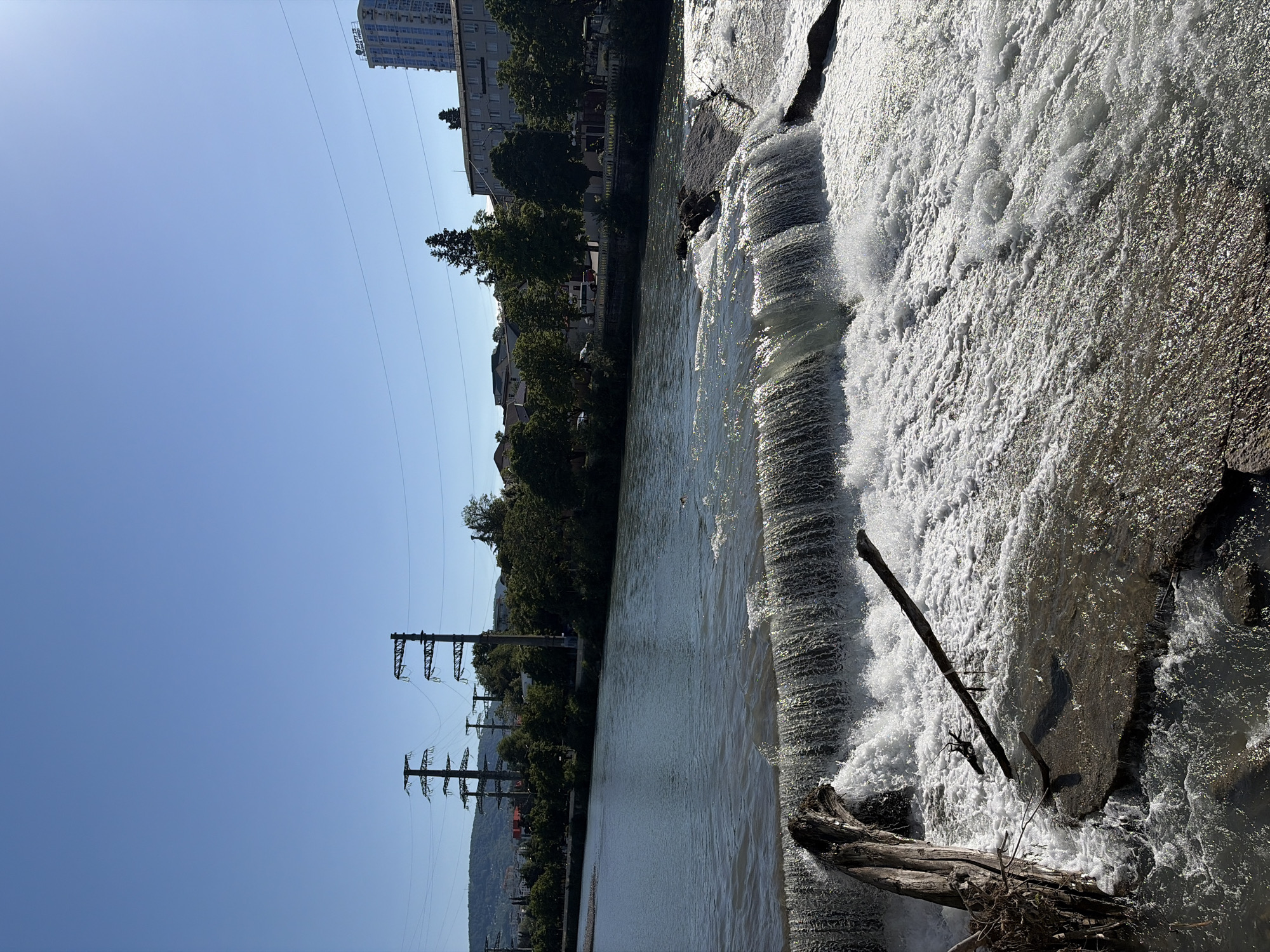 This image captures a vibrant outdoor scene in Sochi, Russia, featuring a wide river with a cascading waterfall or weir, set against a backdrop of urban development and natural landscape.
In the foreground, the river's turbulent waters create a foamy, white cascade as they flow over a rock formation or man-made weir. Several large, dark tree logs and branches are caught in the rushing water at the base of the falls, adding to the rugged texture. The water sparkles intensely under bright sunlight, indicating clear weather.
Across the river, the far bank presents a mix of residential and commercial structures interspersed with lush green trees. A prominent light-colored, multi-story building stands out, and close inspection reveals Chinese characters on a sign, specifically "华为" (Huawei) and possibly "荣耀" (Honor), indicating a commercial presence. Further down the riverbank, a series of tall, dark metal power line towers with multiple overhead wires stretch into the distance, leading towards hazy, verdant mountains that form the horizon.
The sky above is a vast expanse of clear, bright blue, devoid of clouds. The overall impression is one of a bustling urban environment harmoniously coexisting with the powerful natural features of the river and distant mountains. The time of day appears to be midday, given the direct sunlight and absence of long shadows. No people are visible, and the primary activity is the natural flow of the river.