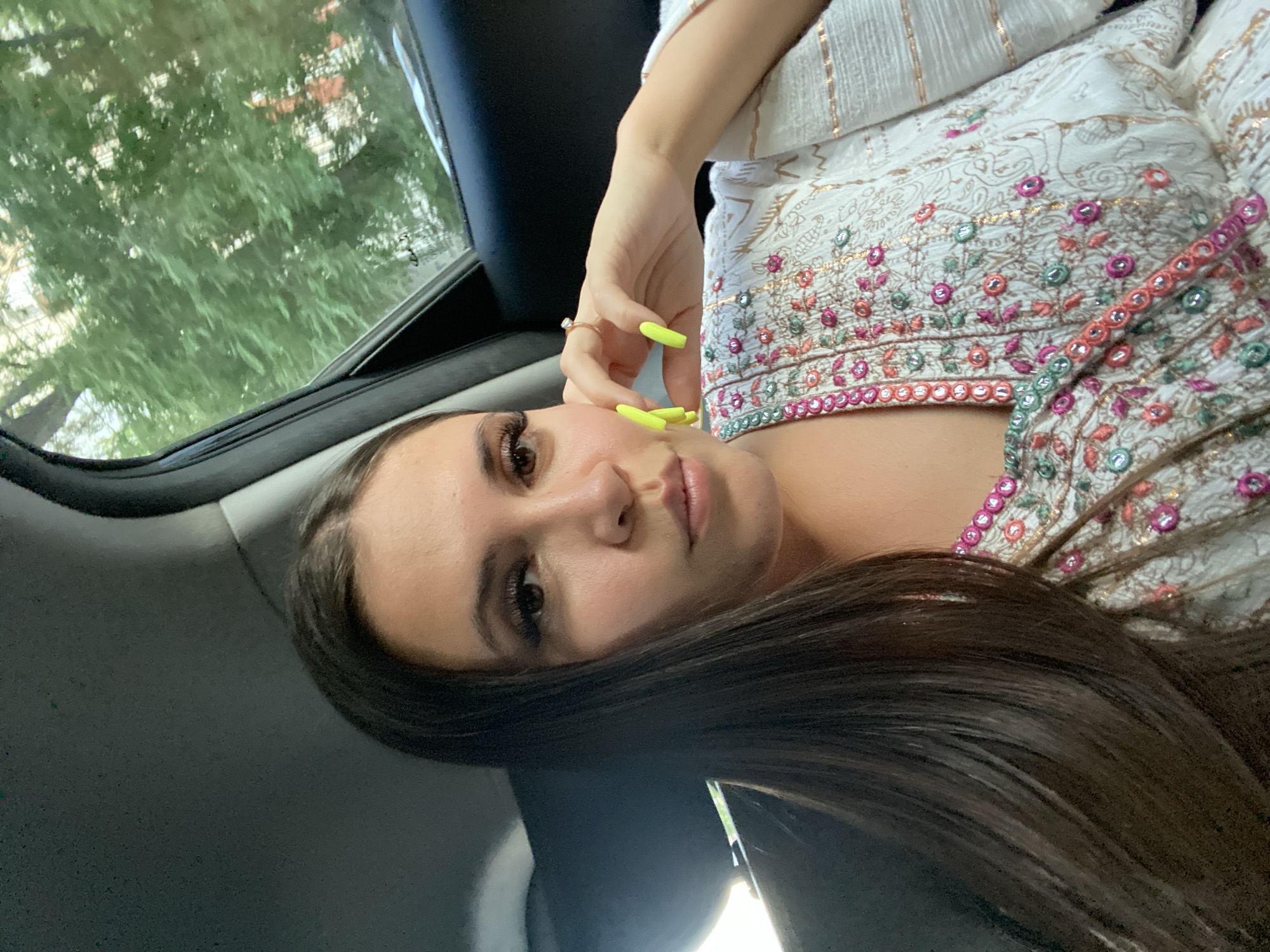 A close-up, horizontally oriented selfie shows a young woman lying down, likely in a car. She has long, dark brown hair that cascades around her face and shoulders. Her eyes are dark and her eyebrows are well-defined with dramatic false eyelashes. Her lips are slightly parted, revealing a subtle smile. She is wearing a white garment adorned with colorful embroidery in shades of pink, green, and gold. Her fingers, painted with bright neon yellow nail polish, are near her face, with one hand resting on her cheek. A thin, gold ring is visible on her ring finger.
The background shows the interior of a vehicle with a dark gray headliner and a window on the left side. Through the window, lush green trees and foliage are visible, suggesting an outdoor setting during the daytime, likely with pleasant weather. The lighting suggests it is daytime, with natural light coming from the window. The overall mood is relaxed and casual, as if captured spontaneously during a car ride. There is no visible text in the image.