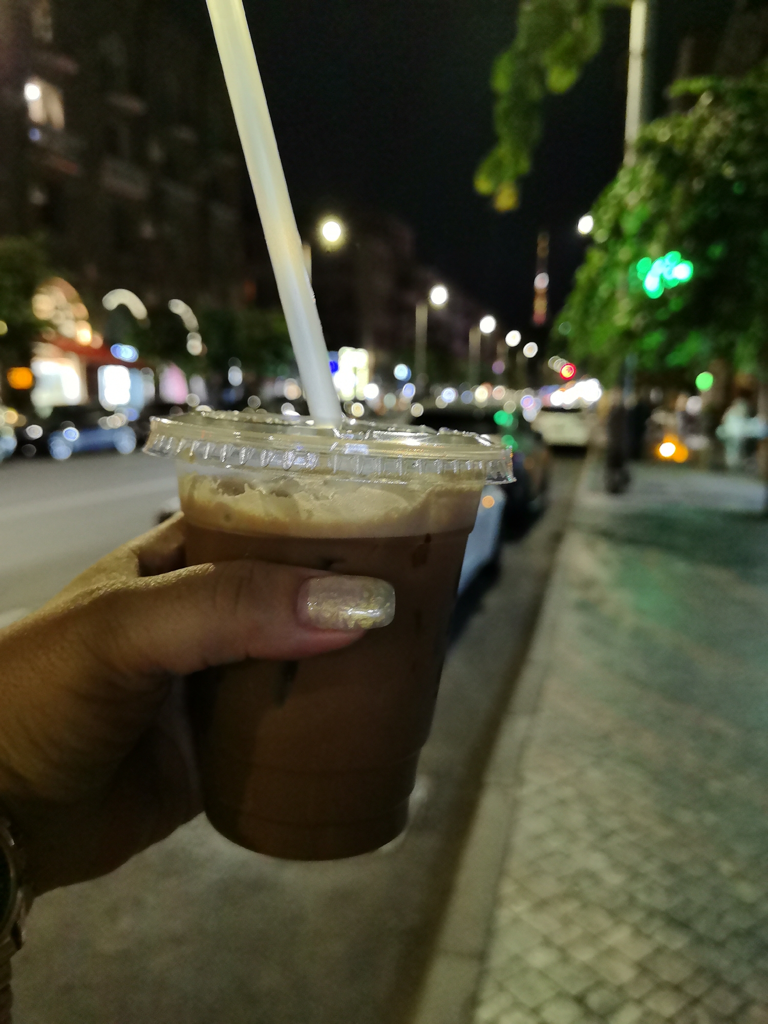 A close-up shot shows a hand holding a plastic cup of iced coffee with a straw, on a street at night. The background is blurred, showing city lights, buildings, trees, and cars, suggestive of a vibrant urban environment. The scene is set in Yerevan, Armenia, likely on a sidewalk adjacent to a street. The hand has metallic nail polish on its fingernails.