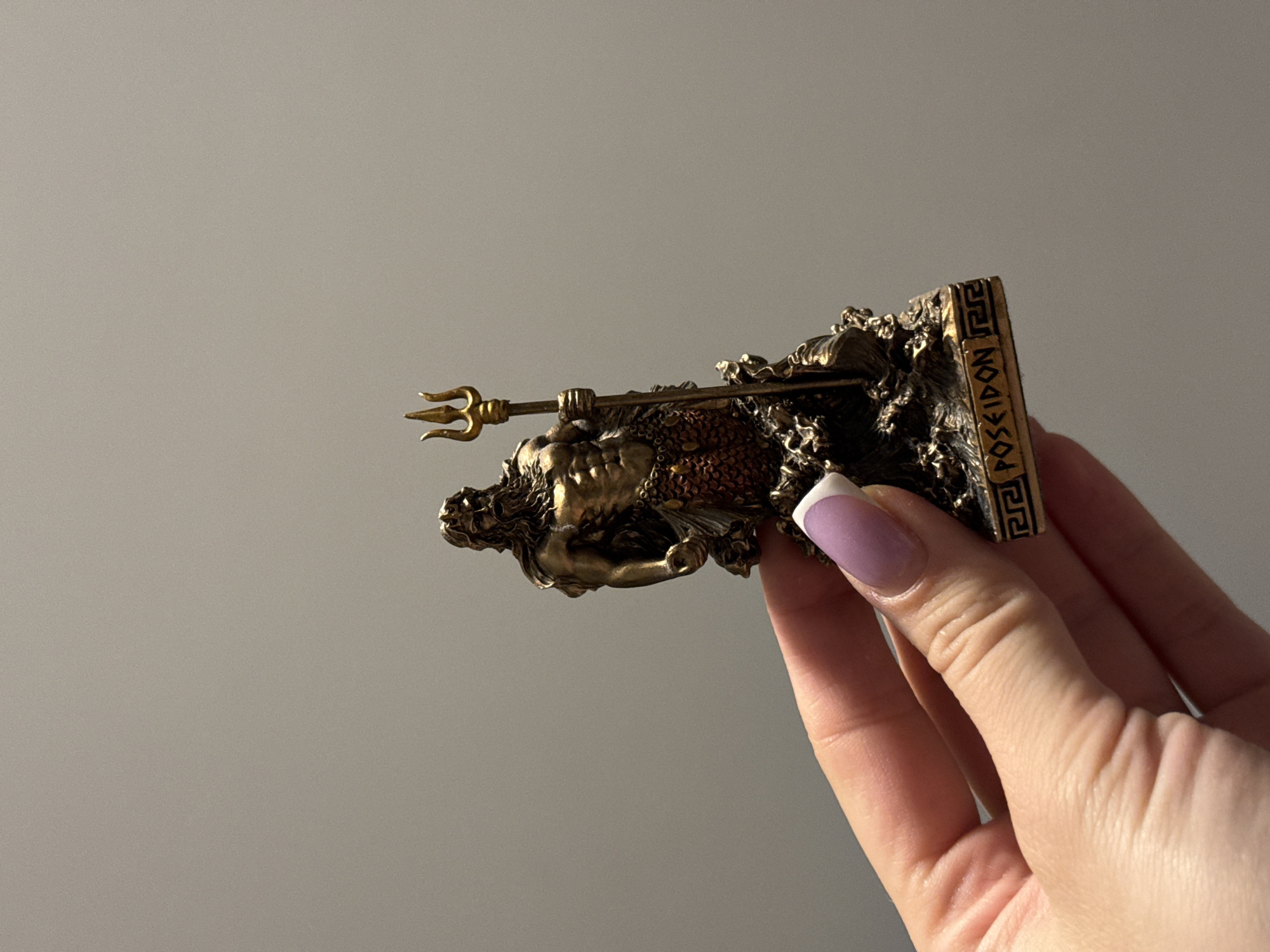A hand is holding a small, bronze-colored figurine of Poseidon, the Greek god of the sea. The figurine depicts Poseidon with a beard, muscular torso, and a trident in his right hand. His lower body appears to be made of scales, suggesting a fish-like tail. The base of the figurine has the word "POSEIDON" inscribed on it, along with a Greek key pattern. The figurine is positioned against a plain, gray background. The image does not contain any information about its location in the city of Łódź, Poland.