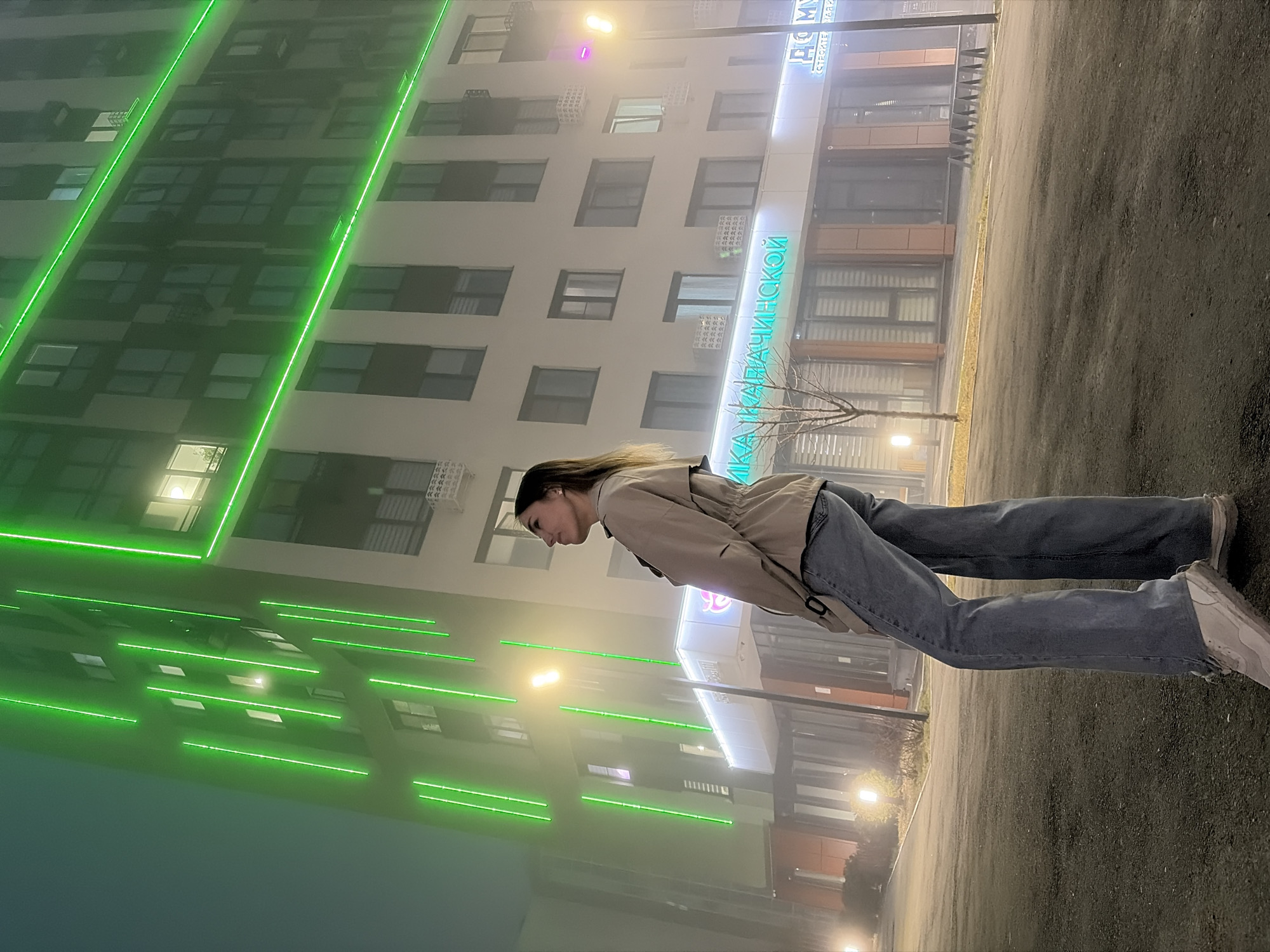 A full-length photograph shows a woman standing in front of a large building. The woman is facing left, wearing blue jeans, a tan jacket, and white sneakers. The building behind her is tall, with multiple floors and numerous windows, adorned with green lights. Signage in Cyrillic script is visible on the building's facade. The location is Izhevsk, Russia.