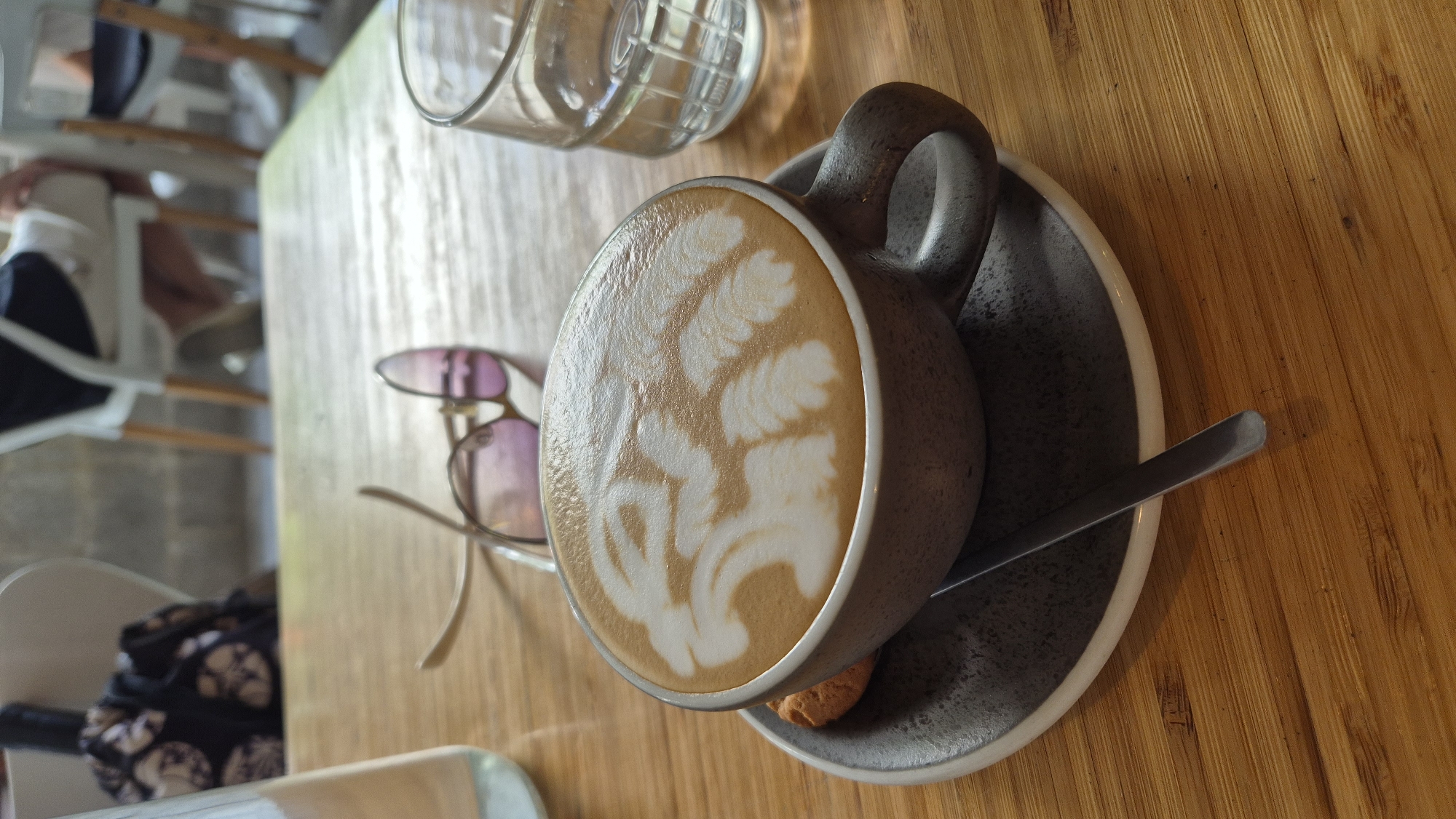 A close-up, eye-level, medium shot captures a cup of latte art on a wooden table in what appears to be a cafe. The latte art depicts a stylized lion's head, with intricate white foam patterns against the brown coffee. The cup is a dark gray, ceramic mug with a speckled finish, resting on a matching saucer. A single, round biscuit, golden brown in color, is placed on the saucer to the left of the cup. A silver spoon, angled downwards, lies on the saucer in front of the cup.
In the background, out of focus, several elements suggest a public setting. A pair of sunglasses with pink lenses and gold frames are placed on the table behind the coffee cup, with their arms splayed. To the right of the cup, a clear glass filled with water is visible. Further back, two individuals are seated at tables, partially obscured. One person, wearing light-colored shorts and a dark top, is seen from the waist down, seated on a white chair with wooden legs. Another person is visible in the far right background, also seated. The environment appears to be an indoor cafe with exposed brick walls and more white chairs. The wooden table has a warm, light brown hue with distinct grain patterns.
The scene is bathed in natural light, casting subtle shadows. The overall impression is one of a relaxed cafe setting, likely in the city of Đà Nẵng, Vietnam, given the typical cafe ambiance and the presence of what could be tourists or locals enjoying a beverage.