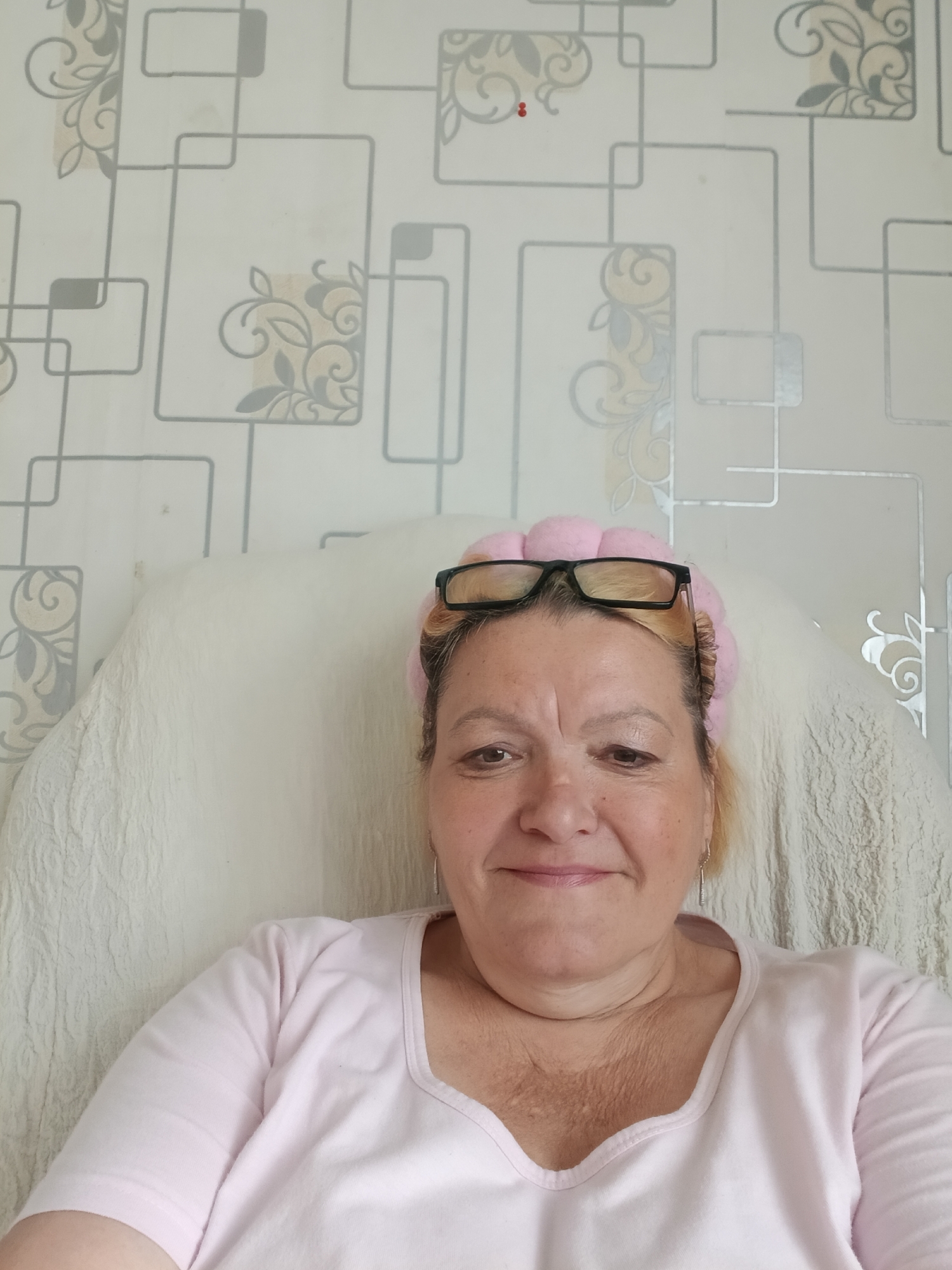 An indoor, head-and-shoulders portrait features an adult woman with light-colored hair, seated and looking directly at the camera with a gentle smile. She is wearing a soft, pink, puffy headband, with black-framed glasses resting on top of it. Her attire consists of a light pink, short-sleeved top with a scoop neck.
The background behind her includes a light-colored, textured fabric, likely a chair cover or blanket, particularly visible behind her right side. The wall behind her is covered in light-toned wallpaper featuring a repeating pattern of interconnected grey geometric outlines (rectangles and squares), interspersed with subtle grey and light beige floral or swirl motifs. The lighting appears bright and even, suggesting it is daytime. The woman has a relaxed and pleasant expression. This media file was captured in Красный Маяк, Russia. There is no visible text in the image.