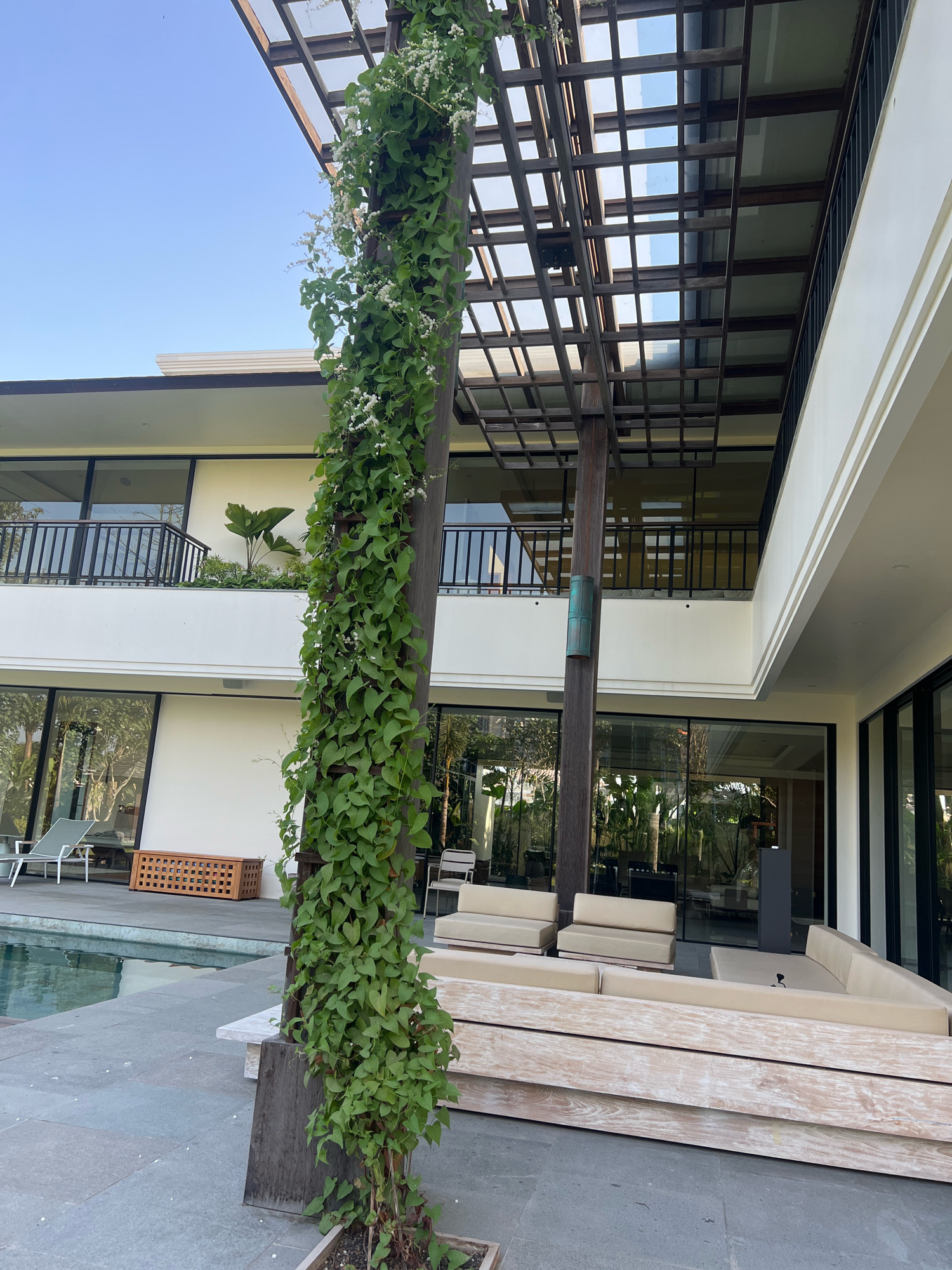 The image shows a modern villa with a swimming pool in the foreground, located in the city of Ngaglik, Indonesia. The villa features clean white walls, large glass windows, and a wooden pergola structure. A lush green vine with small white flowers climbs up a wooden post in the foreground, partially obscuring the view of the villa.
To the left of the vine, a light blue swimming pool is visible, with a lounge chair on the deck. A wooden storage bench with a patterned front sits near the pool. The main structure of the villa has two levels, with balconies on the upper floor. Large glass doors and windows on the ground floor offer glimpses of tropical plants inside.
In the center of the image, an outdoor lounge area is set up with a large, modular sofa made of light-colored wood and beige cushions. The sofa is positioned on a grey tiled patio. The pergola extends over this area, providing shade. The sky is a clear, light blue. The scene depicts a tranquil, luxurious outdoor living space.