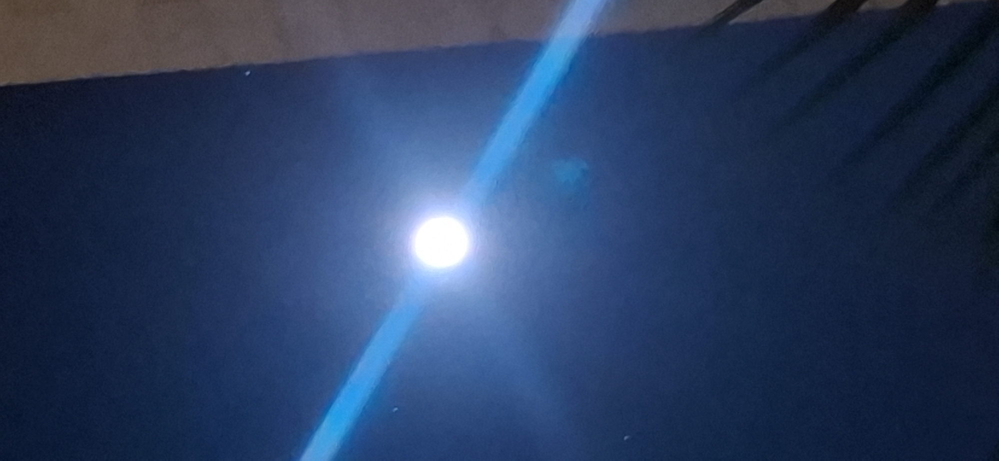 The image depicts a dark, possibly nighttime, scene with a bright circular object, likely the moon, illuminated by a strong light source. A diagonal band of intense blue light cuts across the frame, originating from the left and passing through the bright object. The background is a deep blue, suggesting a night sky. A vertical light brown element is visible on the right edge of the image, possibly a building or wall. In the lower right portion of the image, some dark, shadowy forms that resemble palm fronds or branches are visible. The overall impression is one of artificial light casting a dramatic effect on a dark environment. The image could have been taken in Yerocham, Israel, as it is a city that experiences clear skies suitable for viewing celestial objects.