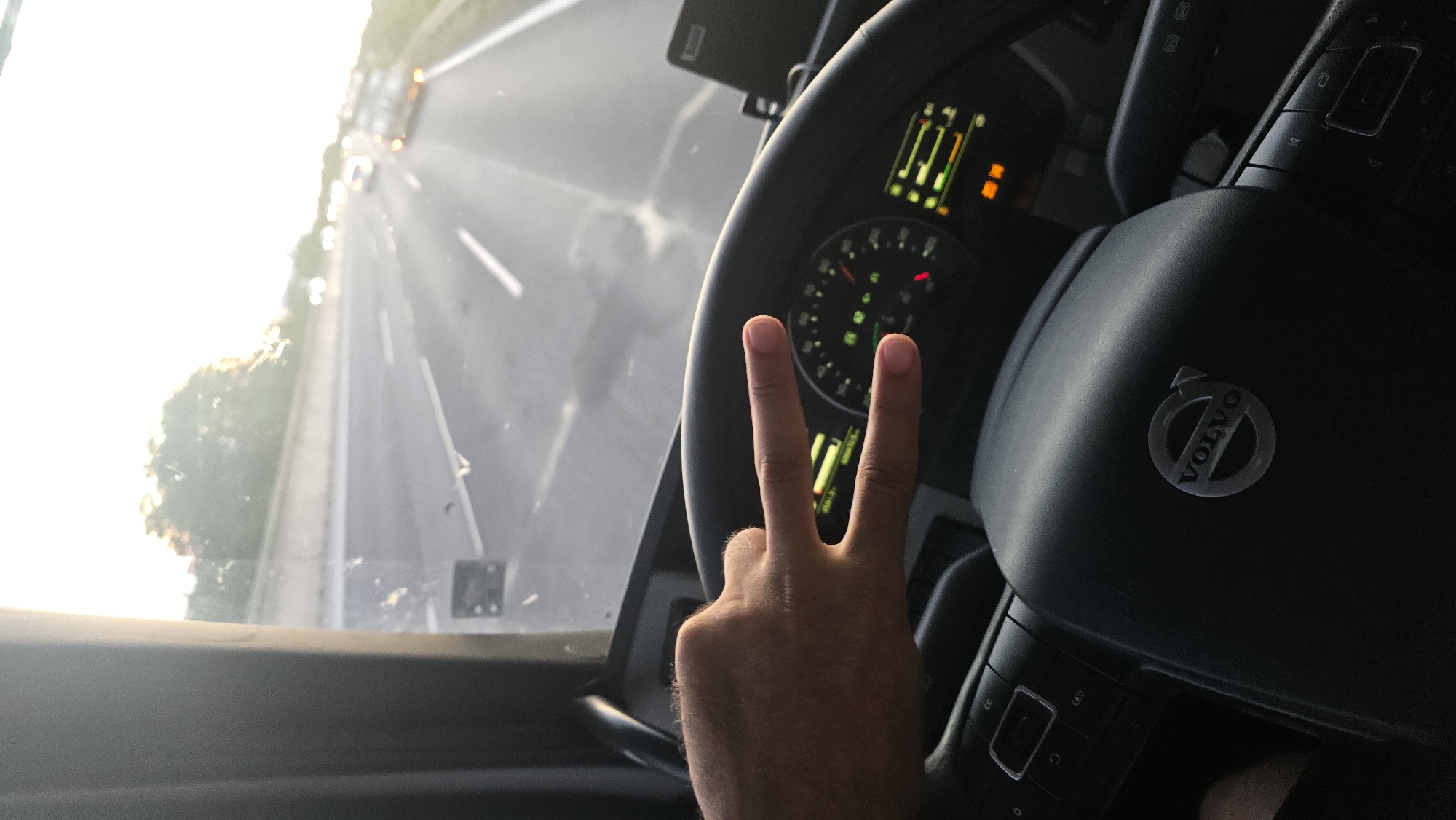 A first-person perspective shot from inside a Volvo truck cab shows the driver's hand making a peace sign. The driver's left hand, with the thumb and index finger extended and the other fingers closed, is positioned in front of the steering wheel. The steering wheel has the Volvo logo in the center and various control buttons. The dashboard displays are visible, with a speedometer and other indicators. Through the windshield, a highway is seen with other vehicles in the distance and trees lining the side. The sky is bright with sunlight, suggesting it is either sunrise or sunset. The image was taken on the D1 highway, specifically in the section near Veľký Biel, Slovakia, a municipality in the Senec District of the Bratislava Region.