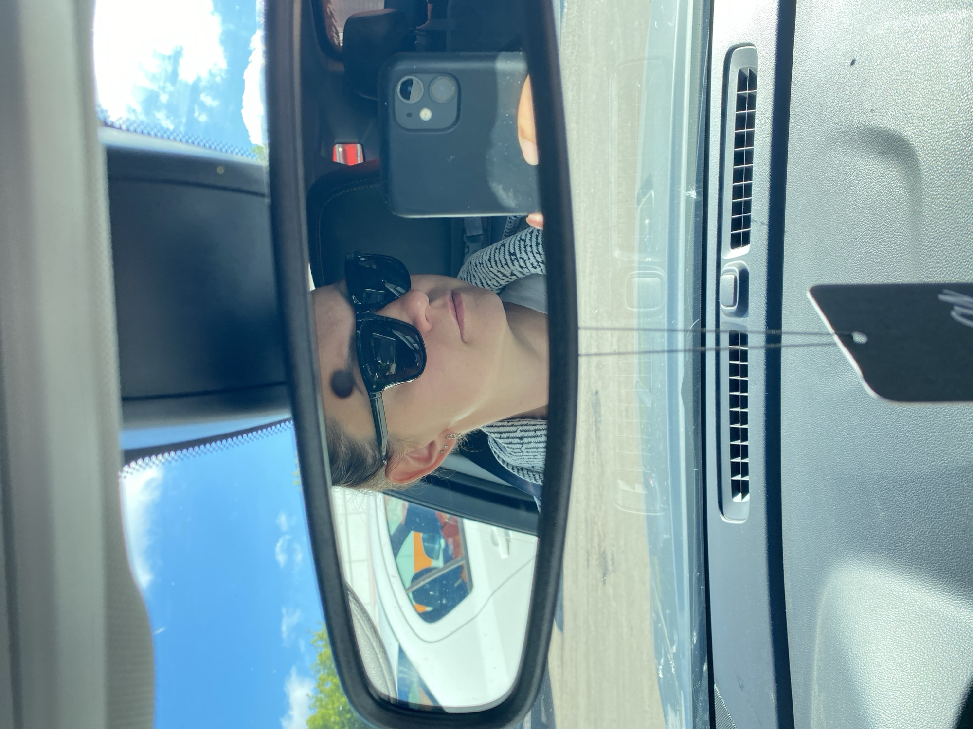 A woman is taking a selfie in the rearview mirror of a car. She is wearing sunglasses and a patterned shirt. The car is parked outside, with other cars and a blue sky with clouds visible in the background. A black air freshener hangs from the rearview mirror. The image is taken from the perspective of the driver's seat, looking forward towards the windshield.