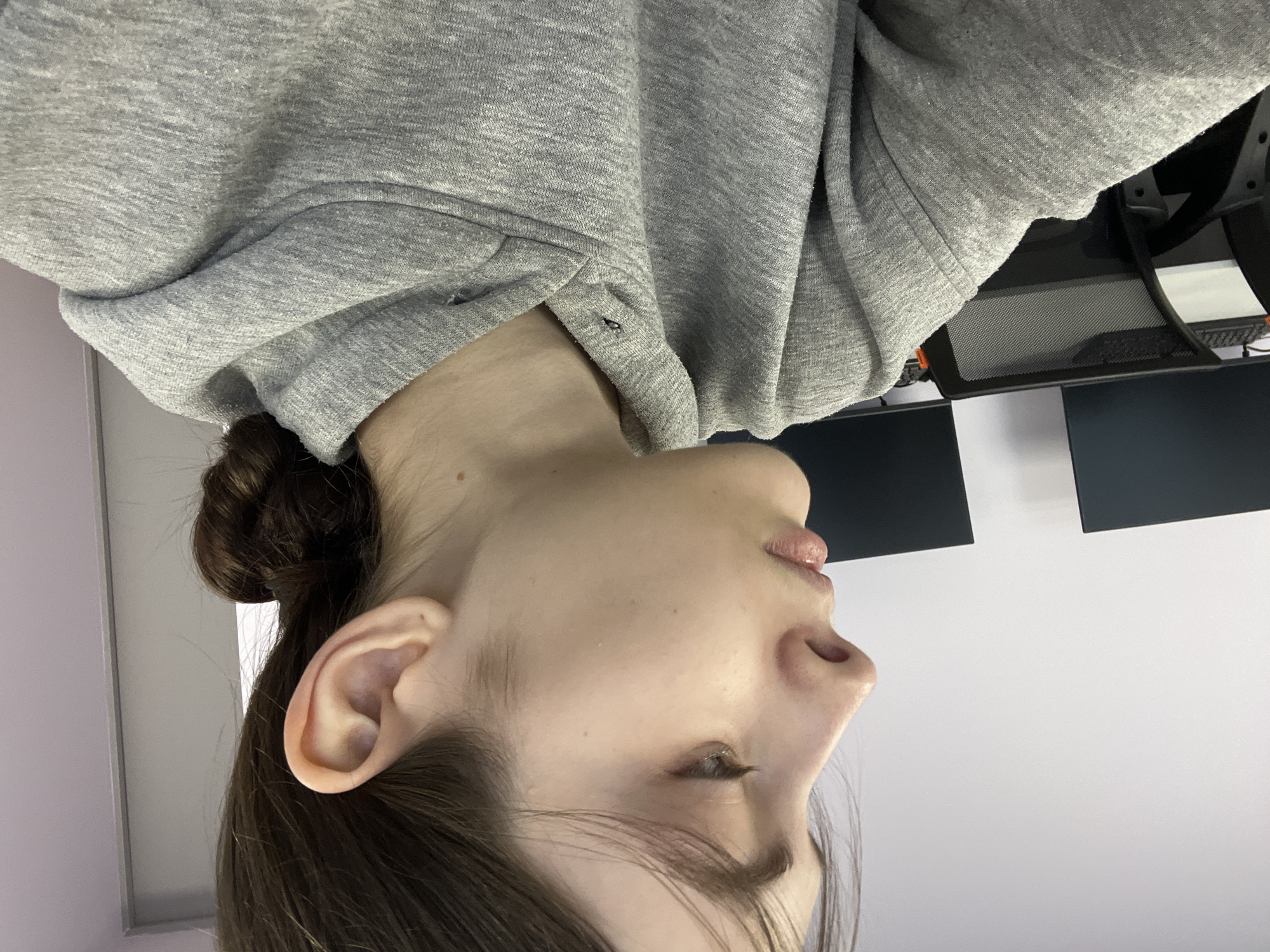 A close-up shot from a low angle depicts a fair-skinned person wearing a gray hoodie. The individual's hair is pulled back into a bun. The subject's face is angled upwards, showing the ear, profile, and nose. The setting appears to be an indoor office environment. Behind the person are dark computer monitors and a white wall. The exact location within Saint Petersburg, Russia, is not discernible from the image alone.