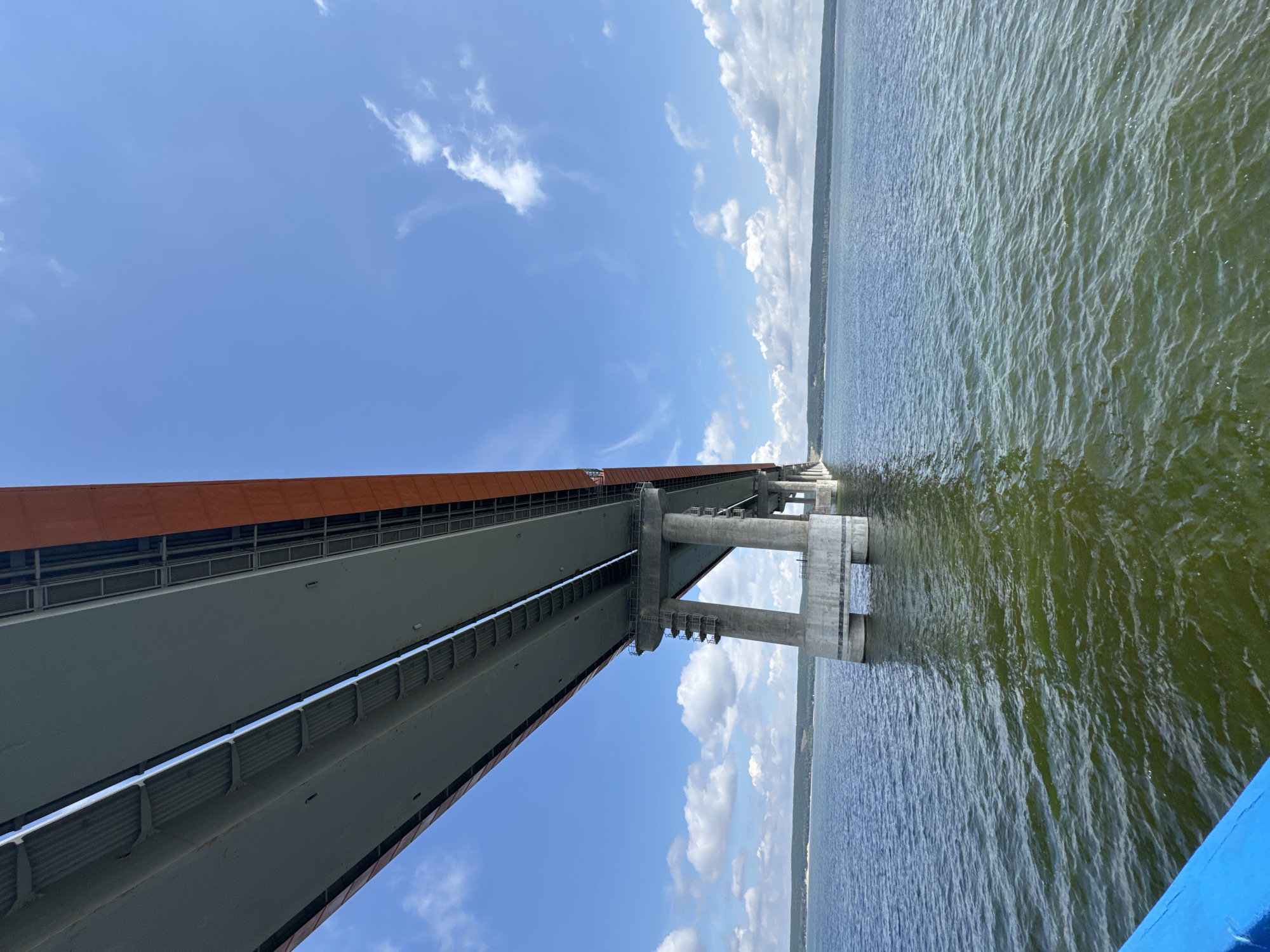 A low-angle shot captures a massive bridge spanning a wide, calm body of water under a bright blue sky dotted with white clouds. The underside of the bridge dominates the upper portion of the frame, revealing its grey concrete deck and orange-painted sides. The bridge is supported by numerous concrete pillars that extend down into the water. The water itself is a deep blue-green, with small ripples disturbing its surface. In the distance, a tree-lined horizon is visible. The scene appears to be taken from a boat, as a sliver of blue railing is visible in the lower left corner. The bridge is part of the M7 Volga Highway over the Sviyaga River near the village of Shelanga, Russia.