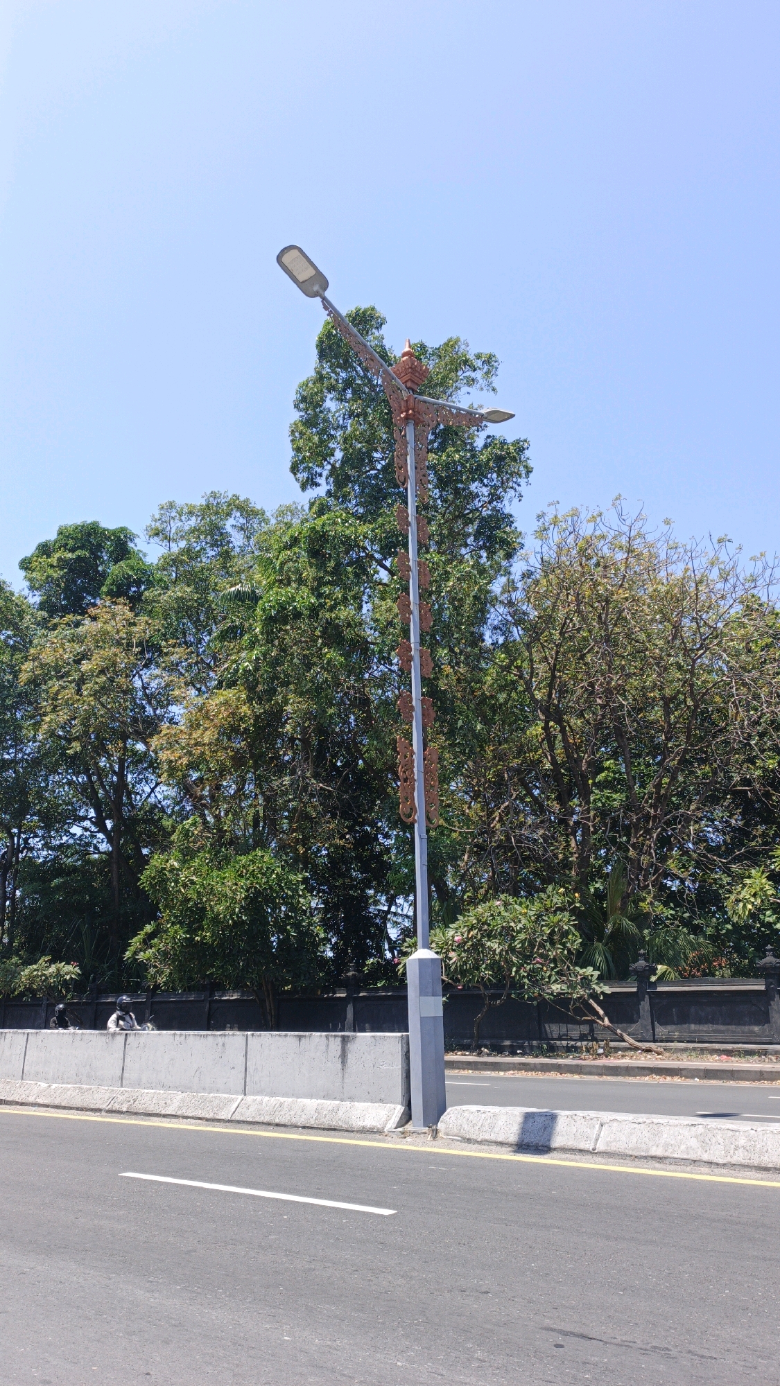 The image shows a decorative lamppost on the side of a road in Denpasar, Indonesia. The lamppost is made of metal and has ornate carvings along its pole. It is topped with two lights and a decorative finial. In the background, there are lush green trees and a dark stone wall. A concrete barrier separates the road from the area behind it, where two people on motorcycles are visible. The road itself is asphalt with white and yellow markings. The sky is clear and blue.