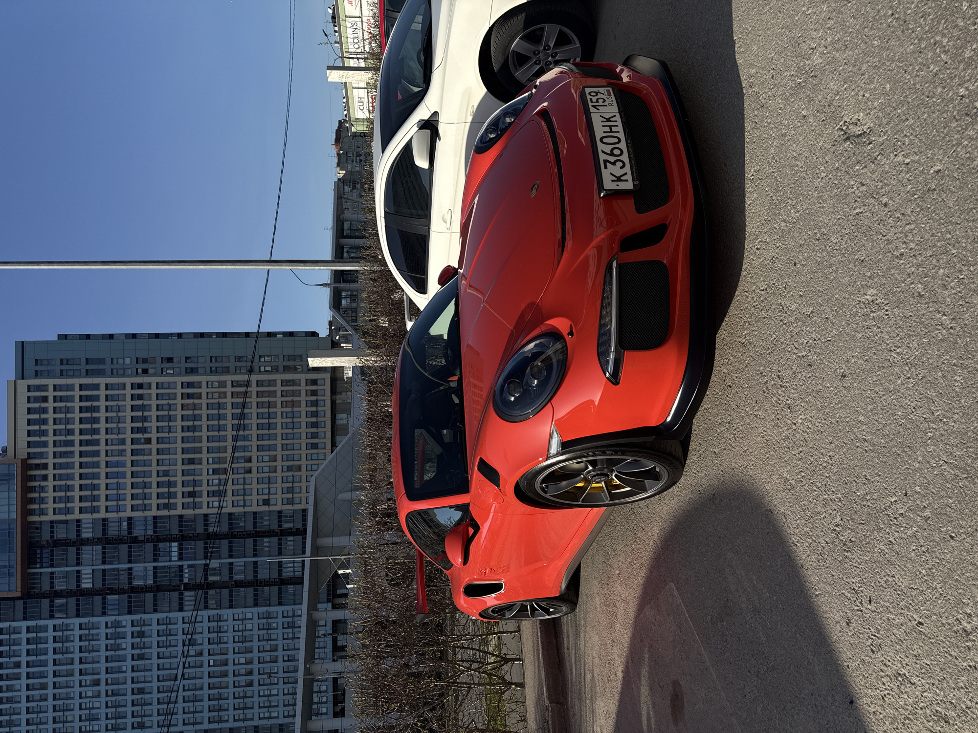 A red Porsche GT3 is parked on a gray asphalt surface. To the left of the Porsche is a white vehicle. A high-rise building is in the background. The license plate on the car reads "K 360 HK 159 RUS". The area is likely in Perm, Russia, based on the license plate.