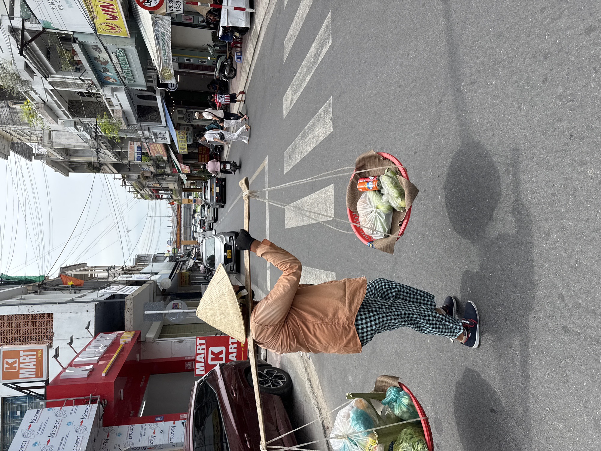 The image captures a street scene in Nha Trang Ward, Vietnam. The focal point is a person wearing a traditional conical hat and a light brown jacket. They are carrying two red baskets suspended from a bamboo pole across their shoulders, likely containing produce for sale. The person is standing on a pedestrian crosswalk on an asphalt street. The surroundings include buildings with shop signs, one prominently displaying "K MART + PLUS." parked cars are visible next to the sidewalk. Overhead power lines crisscross above the street. The lighting suggests it is daytime. A group of people are also using the crosswalk.
