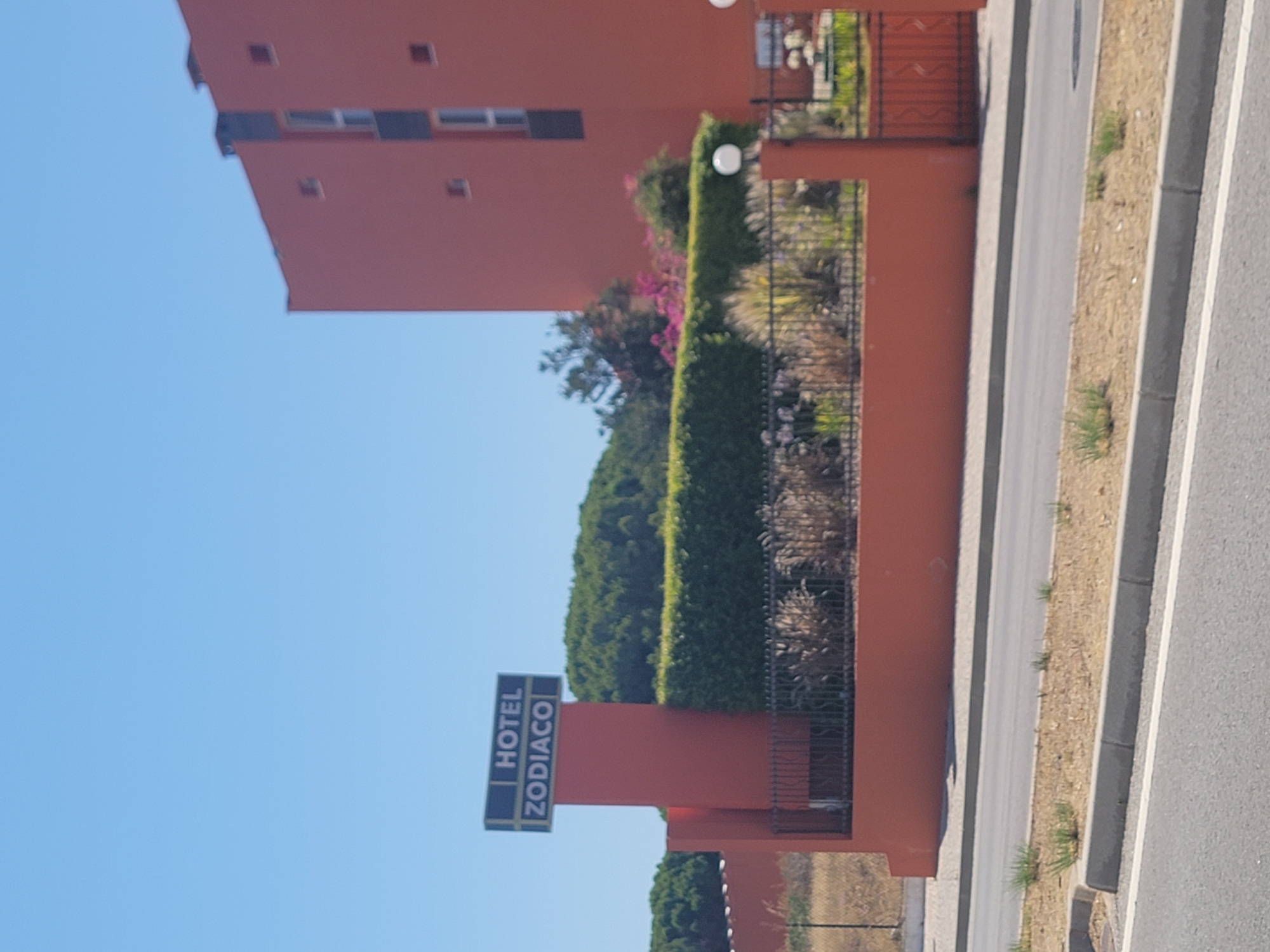 A mid-shot, outdoor, eye-level, daytime, slightly blurry photograph shows the exterior of Hotel Zodiaco in Quarteira, Portugal. The building, painted a bright orange, is partially visible on the right side of the frame. In the foreground, a road and sidewalk are visible, with dry grass and some small plants growing along the roadside. Behind the road, a low orange wall runs across the scene, topped with a hedge. A black metal fence runs along the hedge. A tall, orange pylon stands to the left, supporting a dark rectangular sign that reads "HOTEL" on the top line and "ZODIACO" on the bottom line in white lettering. The sky above is a clear, bright blue. The overall scene depicts the entrance or facade of the hotel on a sunny day.