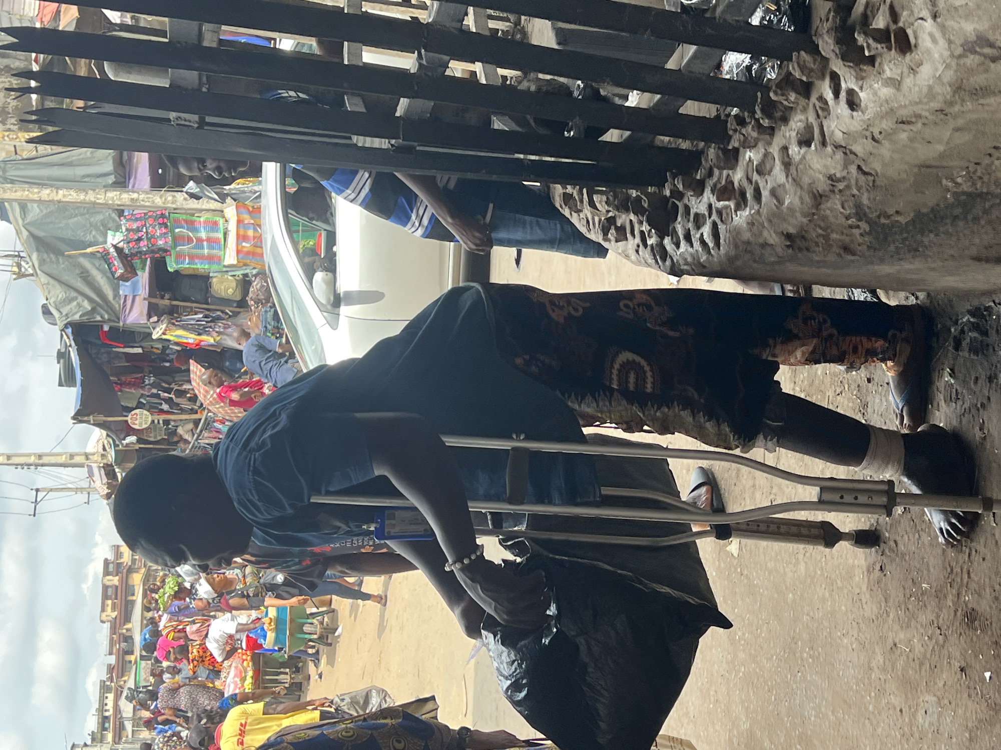 The image depicts a bustling outdoor market scene in Nkpor, Nigeria, likely during the daytime given the bright lighting. The focus is on a person with a disability, using crutches to support themselves. They are dressed in a dark blue t-shirt and a patterned skirt, and a black plastic bag is held in their left hand. The person's right leg appears to be bandaged, and they are wearing a shoe.
The background reveals a busy market with numerous people, stalls, and a white vehicle. Fabrics, bags, and various goods are displayed for sale. The environment appears somewhat chaotic, typical of a busy marketplace. Some overhead structures, possibly made of metal bars, are visible at the top of the image. The ground is a mix of dirt and possibly some concrete. The overall impression is one of everyday life and commerce in a Nigerian market. There are no overt emotional expressions visible from the main subject. The visible text includes "POST" on a sign in the background.
