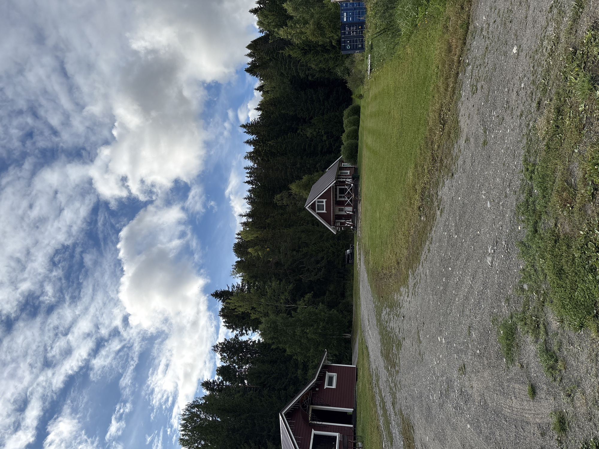 In the city of Rautalakhti, Russia, an outdoor scene unfolds under a partly cloudy sky. A gravel driveway leads into the frame from the left, curving towards a grassy area with a well-maintained lawn. To the left of the driveway sits a small, dark red wooden cabin with a white trim and a white window. Its entrance is open, revealing a darkened interior. Further into the scene, a larger, darker red wooden house with a white trim and a porch is visible, nestled amongst a dense forest of evergreen trees that forms the backdrop. A dark-colored SUV is parked near this house. To the right of the property, two large blue shipping containers are stacked, one on top of the other. The environment is natural, with trees and grass dominating the landscape.