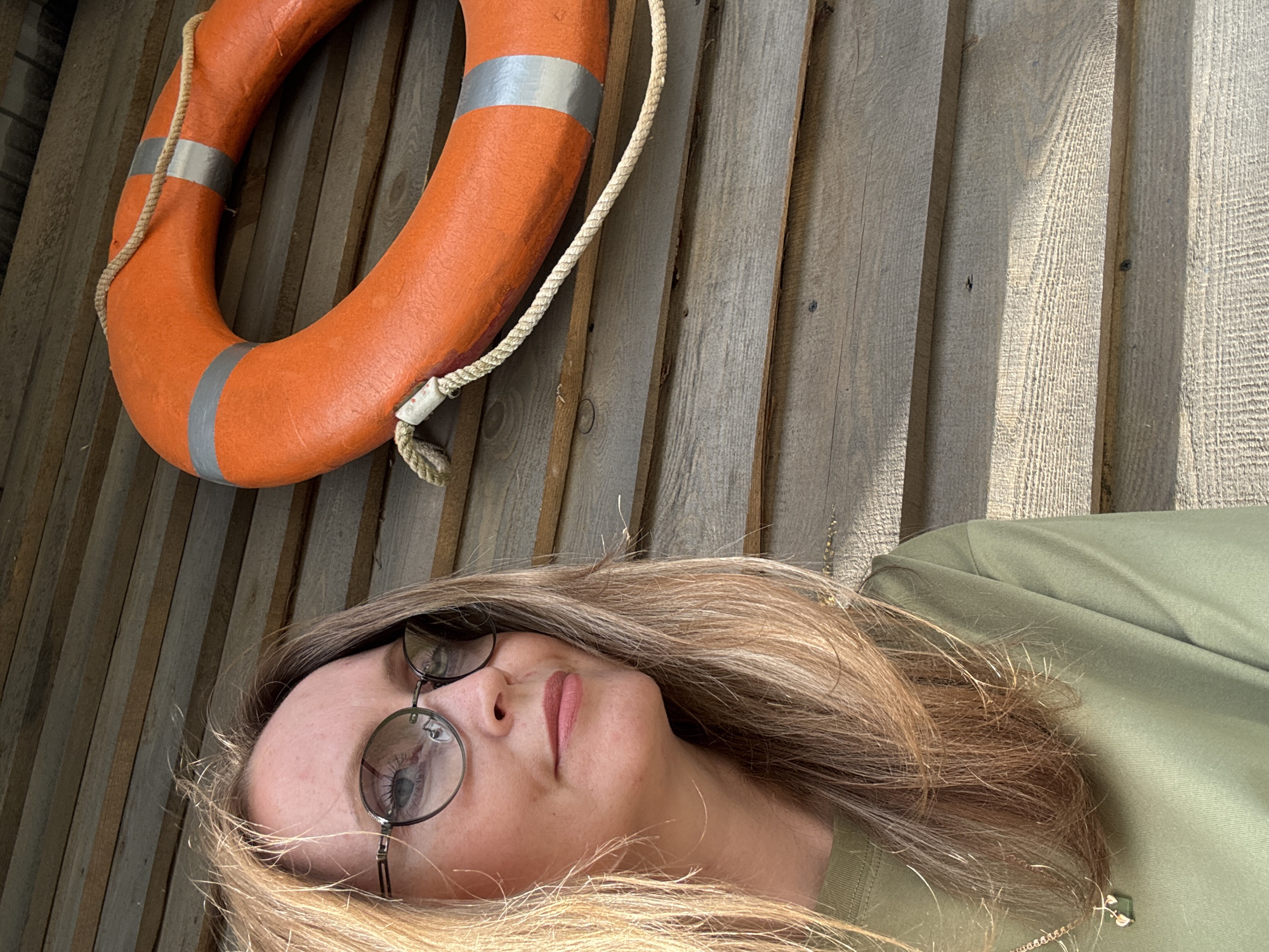 A woman with blonde hair and glasses lies on a wooden surface. She is wearing a green shirt. A life preserver with rope is positioned above her on the wooden surface. The scene appears to be outdoors, possibly near water, given the life preserver. The lighting suggests it is daytime. The woman's expression is neutral. There is no visible text in the image. The wooden planks of the surface are oriented horizontally in the frame.
