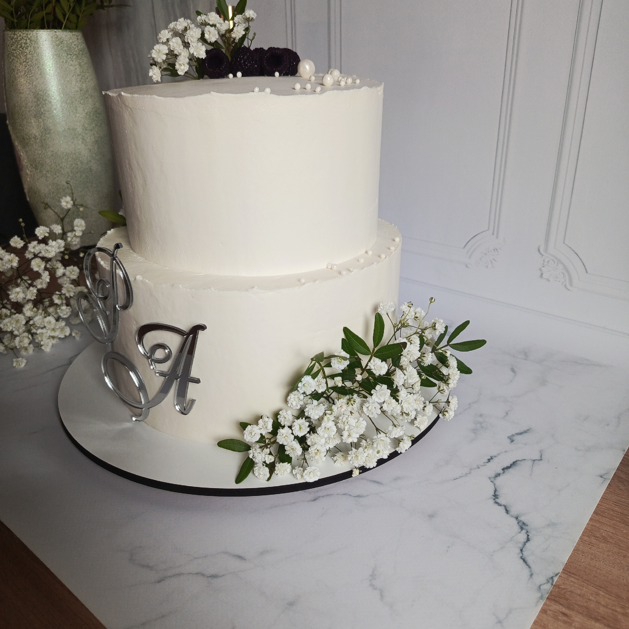 A two-tier white cake, adorned with white frosting, occupies the center of the frame. The smaller top tier features white spherical pearl-like candies, three dark, textured berries resembling blackberries, and a small sprig of white baby's breath flowers. The larger bottom tier displays two reflective, silver-toned initial letters, "J" and "A," in a cursive font, standing upright on its side. A substantial cluster of white baby's breath flowers with green leaves is positioned on the side of the bottom tier. The cake rests on a white circular cake board with a thin black edge. This assembly is situated on a surface resembling white marble with grey veining, which transitions to a light brown wooden surface in the lower right. In the background, a tall, iridescent light green vase containing green foliage is partially visible to the left. Further left, another cluster of white baby's breath flowers is present near the base of the vase. The background wall is light grey or white with raised decorative paneling. This scene is observed in the city of Марковский, Russia.