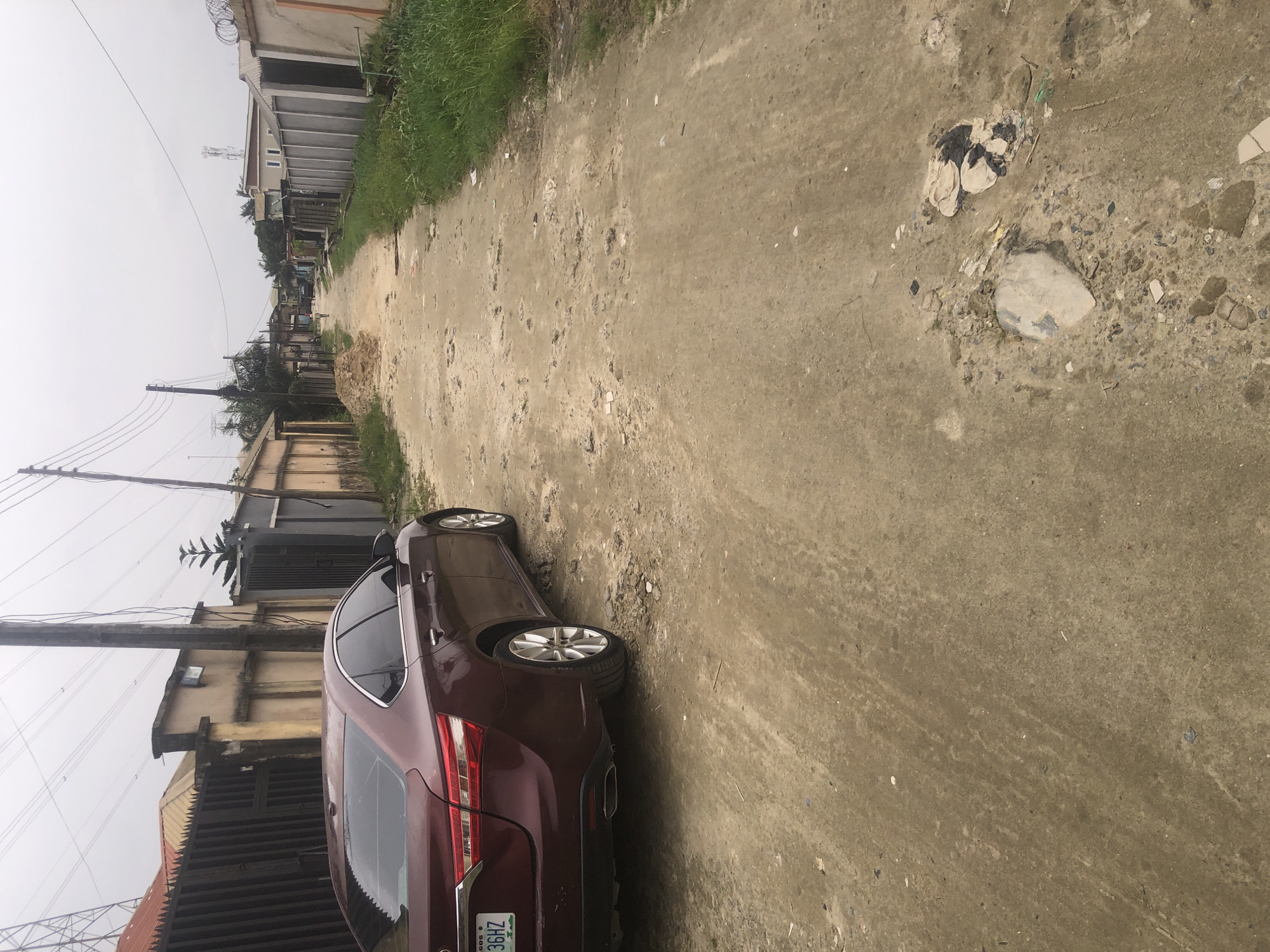 A maroon sedan is parked on a dusty, unpaved road in a densely populated urban area, possibly Ado, Nigeria. The car is positioned on the left side of the road, taking up a significant portion of the foreground. The road itself is uneven and appears to be made of compacted dirt and gravel, with scattered debris and patches of overgrown grass along the sides.
In the background, a row of houses and walls line the street. The architecture suggests a mix of residential and possibly commercial buildings, with varying styles and materials. Fences and gates are prominent features, indicating a sense of security or demarcation between properties. Overhead, power lines crisscross the sky, a common sight in urban Nigerian landscapes.
The overall atmosphere is somewhat overcast, with a muted color palette. The scene depicts a typical street in a developing urban environment, characterized by its unpaved infrastructure and the presence of residential structures. No people are clearly visible interacting with the car or in the immediate vicinity. The image captures a moment of stillness in a potentially bustling urban setting.