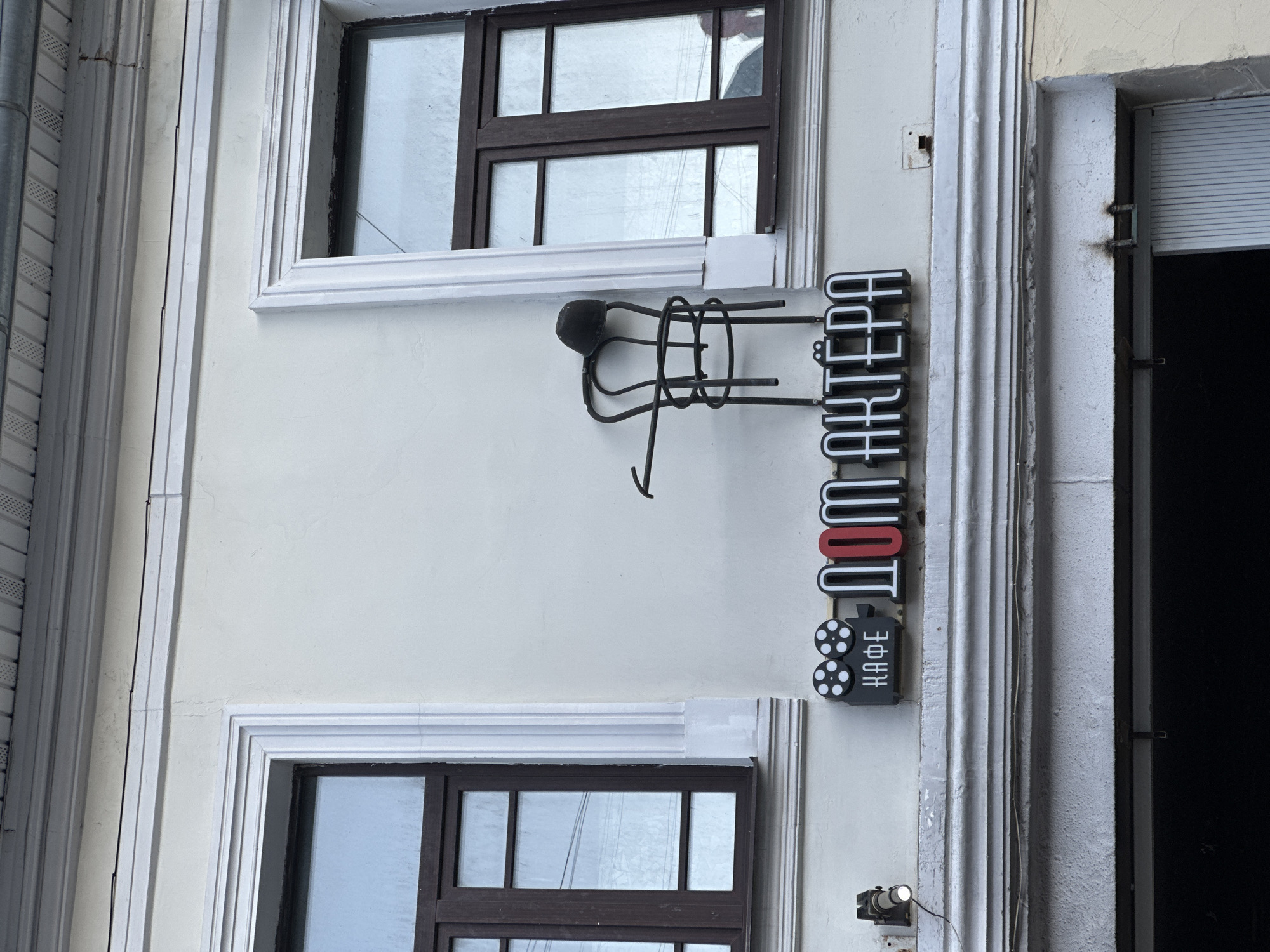 The image shows the facade of a building, likely a cafe, with two windows and a sign. The sign reads "КАФЕ ДОМ АКТЁРА" in stylized lettering, with a film reel icon to the left of "КАФЕ". Above the sign, an artistic metal sculpture of a chair with a hat on it is mounted on the wall. The building is painted in a light, neutral color. The scene appears to be outdoors, with overhead wires visible near the windows. No people are present in the image. The location is identified as Yazvitsevo, Russia.
