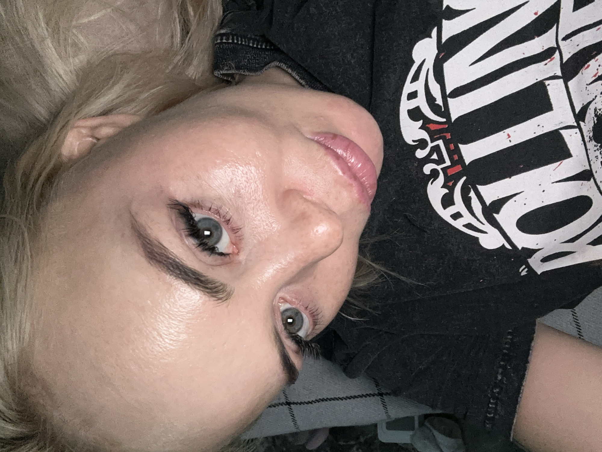 The image captures a close-up, high-angle view of a person, likely female, with blonde hair, lying down and looking directly into the camera. The subject's head is tilted to the left, indicating a relaxed posture, possibly on a bed or couch.
The person has striking light-colored eyes, appearing blue or grey, accentuated by dark eyelashes, which could be mascara or false lashes. Her eyebrows are well-defined and dark. Her lips are slightly parted and appear to have a glossy, light pink or nude finish. Her skin shows a subtle sheen. Her blonde hair is visible around her head and upper left quadrant of the frame, appearing somewhat tousled.
She is wearing a dark grey or black t-shirt with a large white graphic design. Prominent white letters spelling "LLIN" are visible, surrounded by decorative white elements and subtle red accents, strongly suggesting the iconic "Rolling Stones" band logo. The fabric of the t-shirt has a slightly textured or distressed appearance, particularly around the collar and seams.
In the lower right portion of the image, and behind the subject, a checkered or patterned fabric, in shades of dark grey and white, is visible. This fabric likely belongs to a blanket, duvet, or pillow, indicating an indoor setting. The lighting appears to be artificial, suggesting an evening or night-time shot. The overall mood conveyed is casual and relaxed.