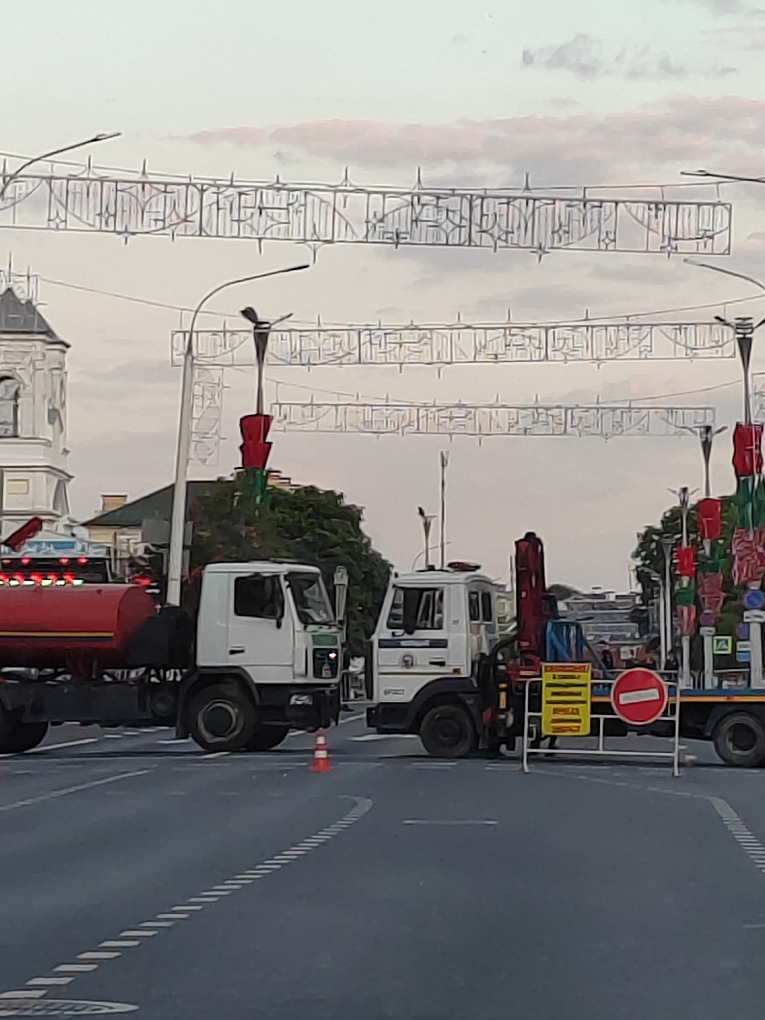 Two white trucks are parked on a paved road in the city of Brest, Belarus. The truck on the left is a tanker truck with a red tank and lights. The truck on the right is a utility truck with a crane. A barricade with a "no entry" sign is placed between the trucks. Decorative lights are strung across the road overhead, and trees and buildings are visible in the background. The sky is overcast.