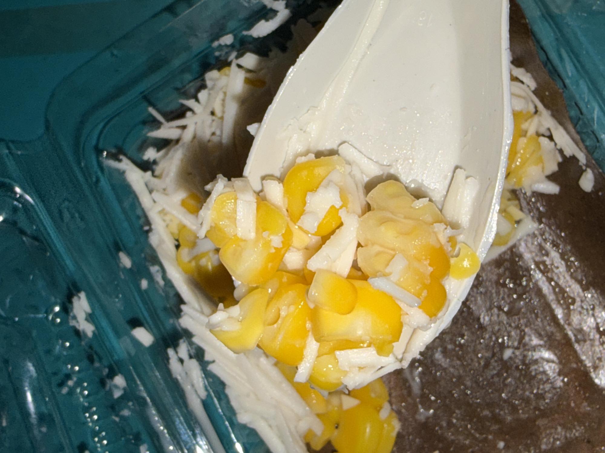 A close-up shot shows a white plastic spoon filled with corn and shredded white substance, possibly cheese or coconut, against a dark brown textured background. The spoon is positioned over a clear plastic container with a teal-colored lid and sides. The container appears to hold more of the corn and shredded white substance. The lighting is direct and harsh, creating bright highlights on the food and spoon, and casting shadows within the container. The scene is indoors, with no view of the external environment or city of Tupah, Indonesia.