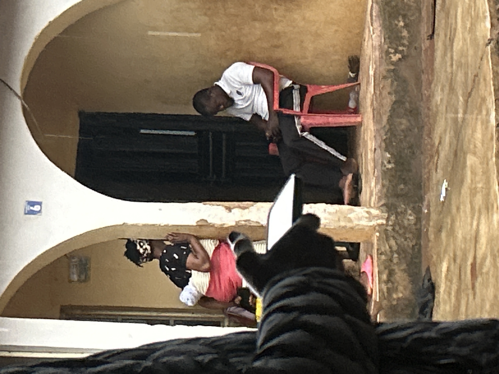 This is a full, eye-level shot of a weathered building in Ikorodu, Nigeria, with an arched entryway leading to a dark doorway. In the foreground, the blurry, dark-clothed arm and hand of a person holding a white object extend into the frame from the left. Inside the entryway, to the left, a woman wearing a black and white patterned headscarf, a black shirt with white dots, and a pink skirt is seen from the back, holding a baby wrapped in a light-colored cloth. Her hands are pressed against the wall. To the right, a Black man wearing a white polo shirt and dark pants sits slumped in a bright pink plastic chair, looking down at what appears to be a mobile phone in his hands. He is positioned near the doorway. The building's facade is textured and appears aged, with peeling paint or plaster. The ground in front of the building is uneven and damp, with some debris visible, including a pair of pink flip-flops. A number "6" is visible on a blue sign above the woman's head. The overall impression is a snapshot of everyday life in a somewhat rustic urban setting.