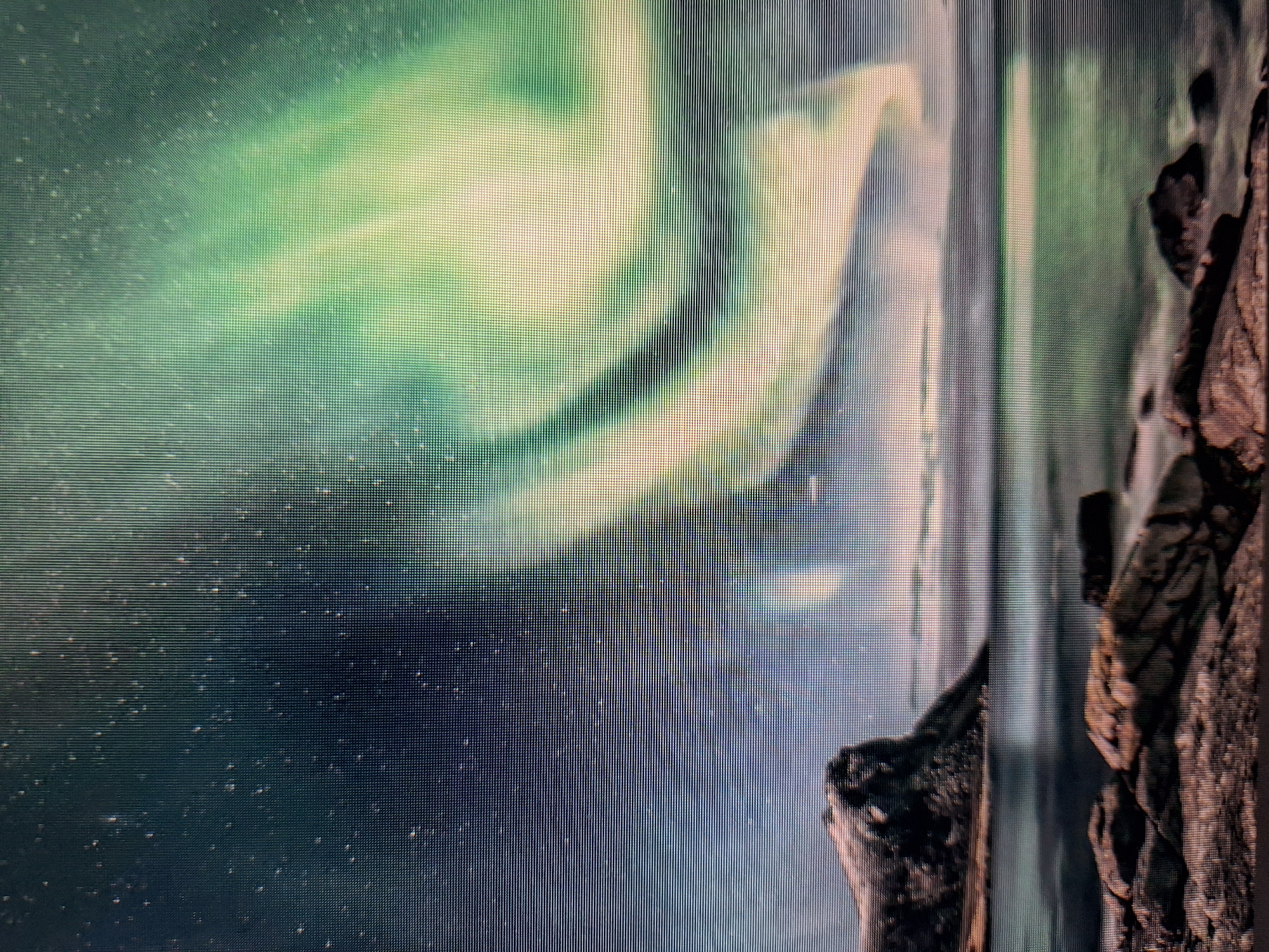 This is a photograph of the Aurora Borealis over a coastal landscape. The sky is dark and filled with stars, and large, swirling ribbons of green and white light from the aurora dominate the upper portion of the image. Below the sky, a calm body of water reflects the lights from the aurora. On the left side of the image, a rocky, snow-capped mountain juts out into the water. In the foreground, large rocks are visible along the shoreline, with the water lapping at them. The scene is a natural landscape, likely in a northern latitude known for aurora sightings. There are no people, buildings, or any urban elements present, and therefore no connection to the city of Varna, Bulgaria, can be inferred from the image.