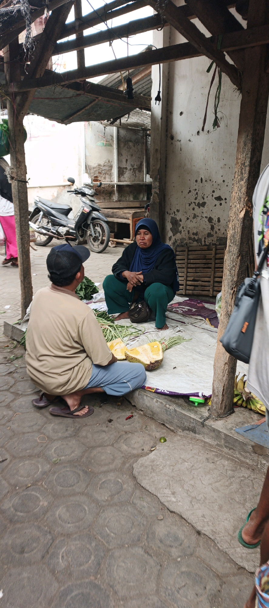 In Kasiyan, Indonesia, a street scene unfolds under a dilapidated wooden shelter. Two individuals are engaged in what appears to be a market transaction. A man, facing away from the camera and wearing a beige t-shirt, a blue cap, and flip-flops, is crouched on the patterned concrete ground. He is positioned to the left of a woman who is also crouched, facing the man. The woman is wearing a blue hijab and green trousers, and her hands are clasped in her lap.
Between them, spread on a light-colored tarp with a purple border, are various items for sale. A halved jackfruit is prominently displayed, its yellow flesh exposed. A bunch of green, long vegetables, possibly beans or a similar produce, lies next to it. In the background, a black motorcycle is parked. The environment suggests a simple, open-air market stall, with weathered walls and a rough, uneven ground surface. The overall impression is one of everyday life in a local Indonesian community.