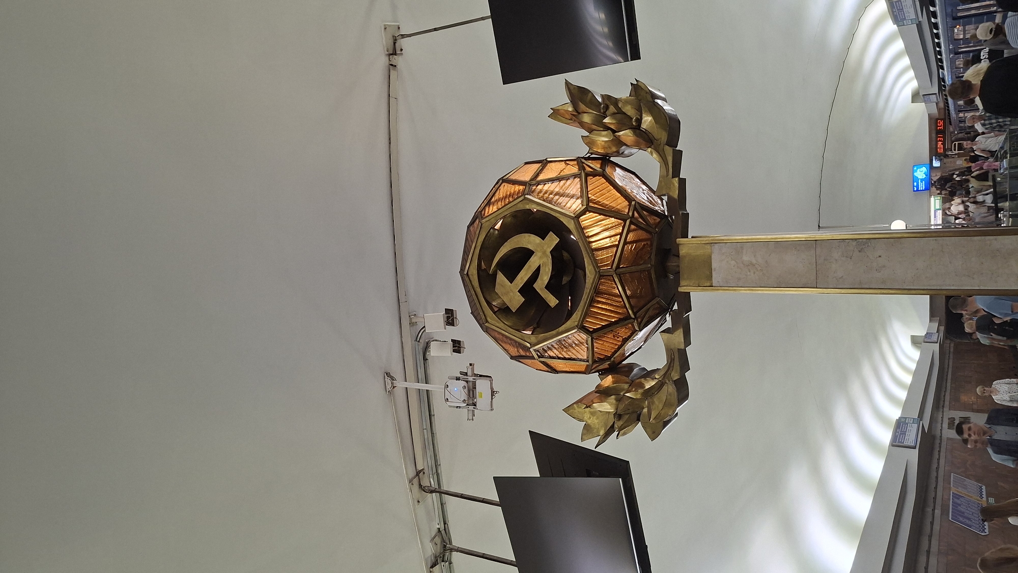 This image depicts a view of the platform at the Ploshcha Peramohi metro station in Minsk, Belarus. Dominating the center of the frame is a large, ornate light fixture with a spherical, amber-colored glass shade encasing a golden hammer and sickle emblem, a symbol of the Soviet Union. The fixture is supported by a tall, rectangular column with marble-like accents and gilded detailing. On either side of the fixture, gold-colored laurel wreaths extend outwards. Above, suspended from the ceiling, are various cables and what appear to be two modern surveillance cameras and two dark, flat-screen monitors. The ceiling itself is white and features distinctive wave-like lighting patterns. In the background, people are visible on the platform and ascending a staircase, indicating the station is in operation. A train car can be seen on the right side of the image, and digital signs displaying times and numbers are present along the platform. The overall environment suggests an indoor public space, specifically a metro station with Soviet-era architectural influences.