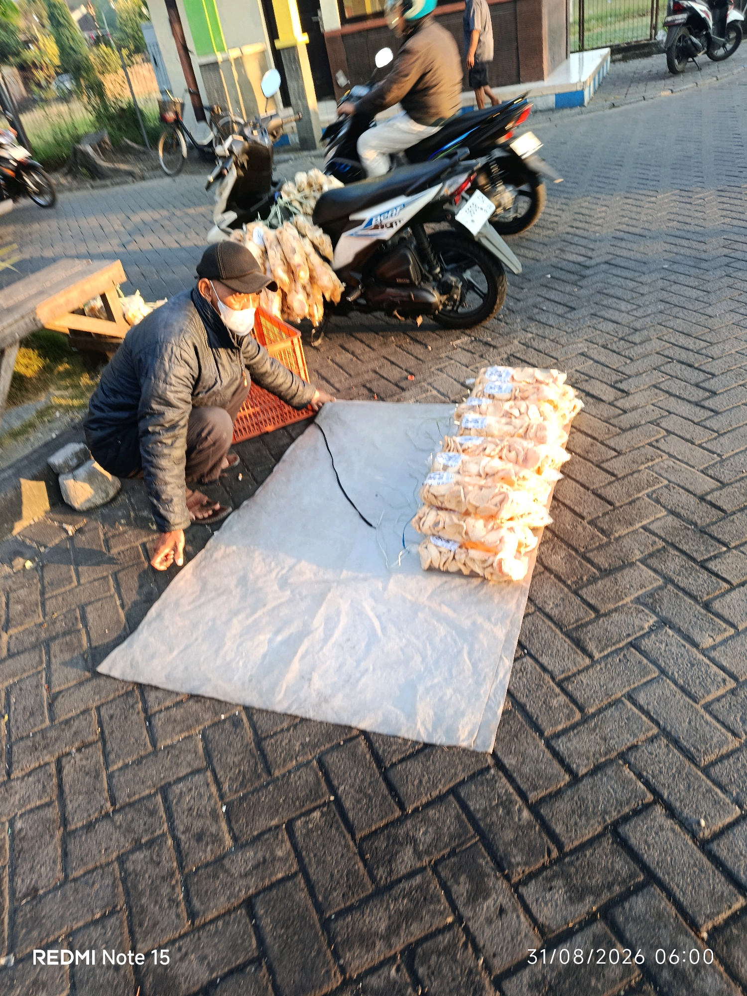 In Keboansikep, Indonesia, a person wearing a black baseball cap, a face mask, and a dark puffer jacket is crouched on a brick-paved surface. They are handling a large white tarp spread on the ground. To the left of the person, an orange plastic crate is visible, containing a pile of food items wrapped in light-colored packaging, possibly smoked meat or sausages. In the background, several motorcycles are parked, some with their lights on, suggesting they are used for transport or as part of a market stall. The scene appears to be outdoors, possibly during early morning or late afternoon given the low angle of the sunlight casting shadows.