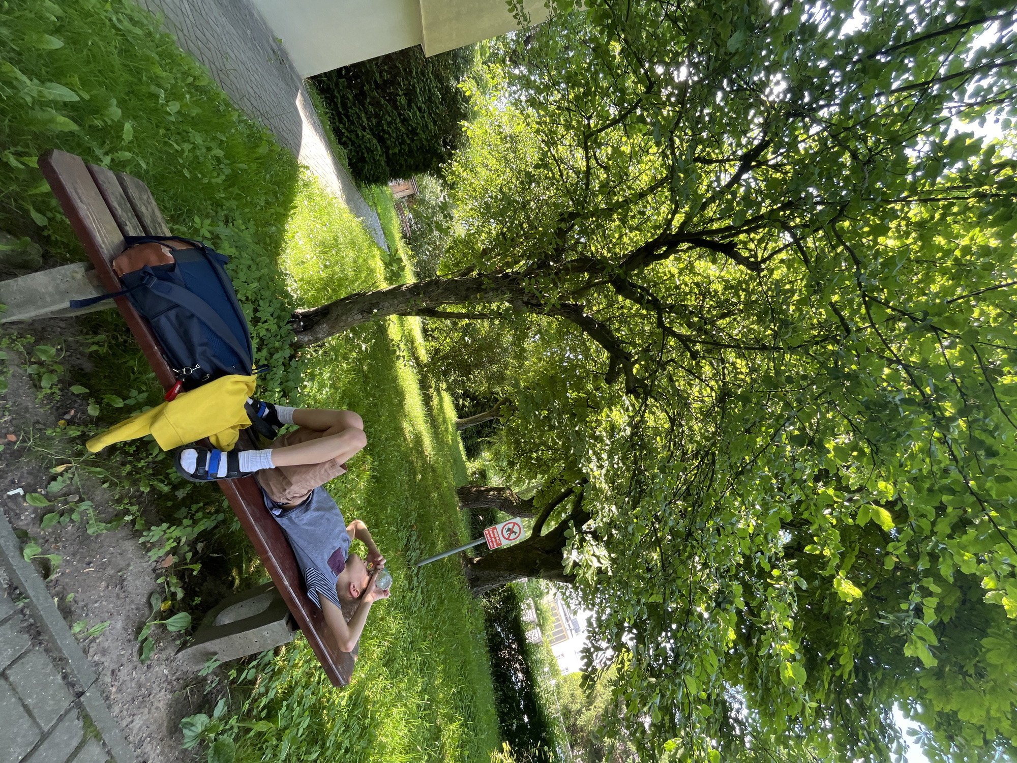 A young boy is lying on a wooden bench under a large tree, with his arms behind his head and a clear plastic bottle held up to his eye. He is wearing a striped t-shirt, shorts, socks, and sandals. A backpack and a yellow bag are on the bench next to him. The bench is in a grassy park, with a paved path and a building visible in the background. A sign with a prohibition symbol is posted on a pole to the right of the bench. The sun is shining through the leaves of the tree, casting dappled shadows on the ground.