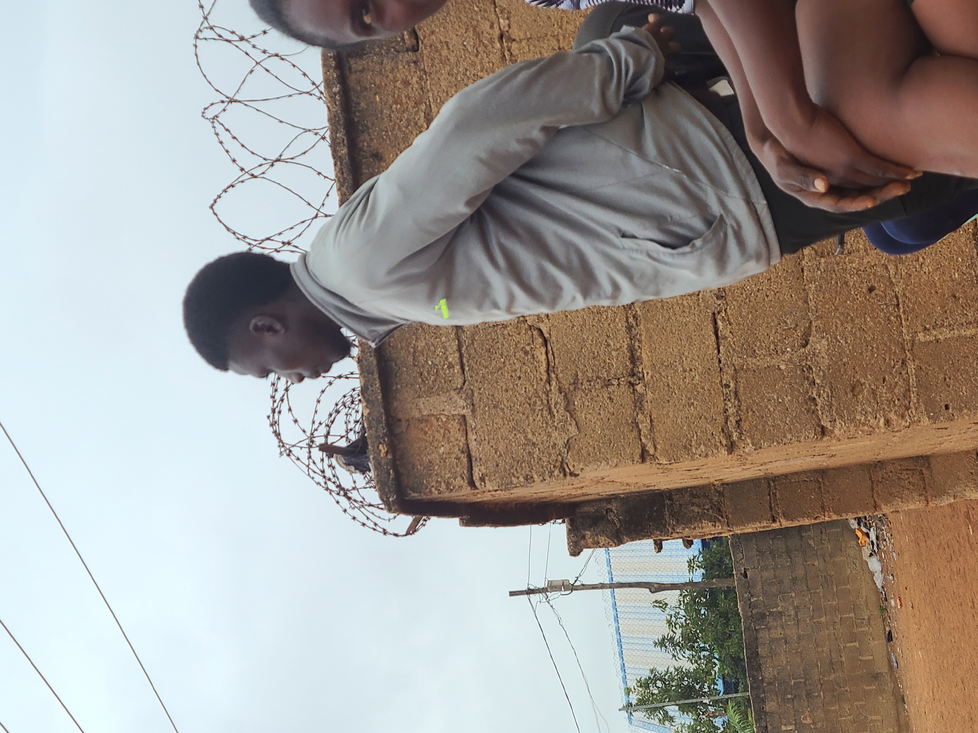 A medium, low-angle, eye-level shot shows two people standing on a brown brick wall topped with barbed wire. One person, a Black male, is seen leaning over the wall, looking down. He is wearing a grey jacket with a small lime green logo on his left side, and has dark hair. The other individual’s face is out of focus, and is standing on the wall behind the man. Below the wall, is a neighborhood scene in Barakin Lerawi, Nigeria, with power lines, vegetation, and a structure. The sky is visible, and looks overcast.