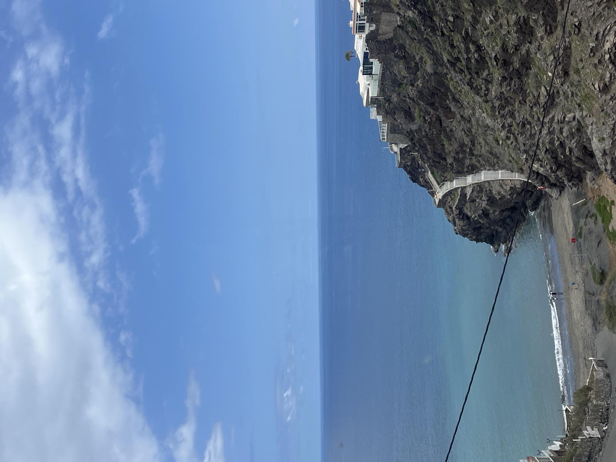 This vertical landscape photograph showcases a stunning coastal scene at San Bartolomé de Tirajana, Spain. On the right, a steep, rugged cliff face, dark and rocky with scattered green vegetation, plunges into the ocean. Several modern, white-washed buildings are precariously perched along the cliffside, some featuring balconies or terraces. A distinctive white walkway with railings snakes along the cliff, connecting various sections and potentially leading towards the beach below.
To the left, the calm, blue-green expanse of the ocean stretches towards the horizon, with gentle waves breaking on a dark sandy or pebbly beach at the base of the cliff. A few small figures are visible on the beach, appearing to be walking near the waterline. A prominent thick black cable or wire diagonally crosses the image from the upper right, over the cliff and beach, and seemingly into the water, suggesting a transport system like a funicular or zipline. Small white structures and a red object (possibly a lifeguard post or sign) are also present on the beach.
The sky is a bright, clear blue with scattered white clouds, ranging from wispy to more defined, indicating a sunny daytime scene. The image captures a dramatic interplay between natural coastal beauty and unique cliffside architecture. There is no visible text in the media file.