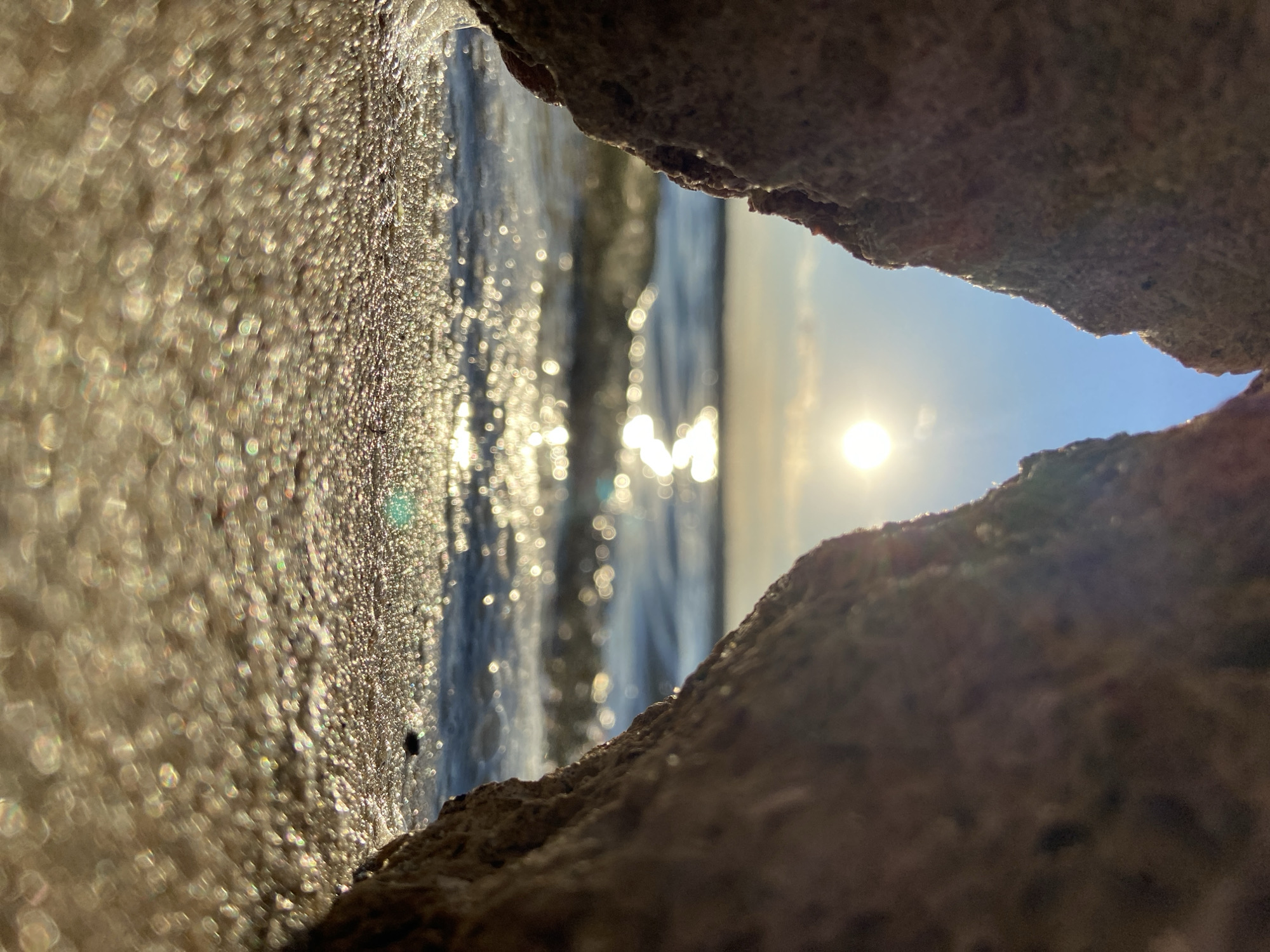 This is a low-angle, close-up, vertical shot of a body of water seen through a gap between two rocks. The water is choppy and reflects the bright sunlight, creating a shimmering effect with numerous bokeh lights. The sun is visible in the upper right portion of the frame, low in the sky, indicating either sunrise or sunset. The sky is a clear, pale blue. The rocks framing the scene are rough and textured, with one on the left appearing wet and glistening with small droplets, possibly from the water. The other rock is on the right, forming the other edge of the gap. The overall mood is serene and peaceful, enhanced by the natural elements and the warm light. There is no visible text in the image.
