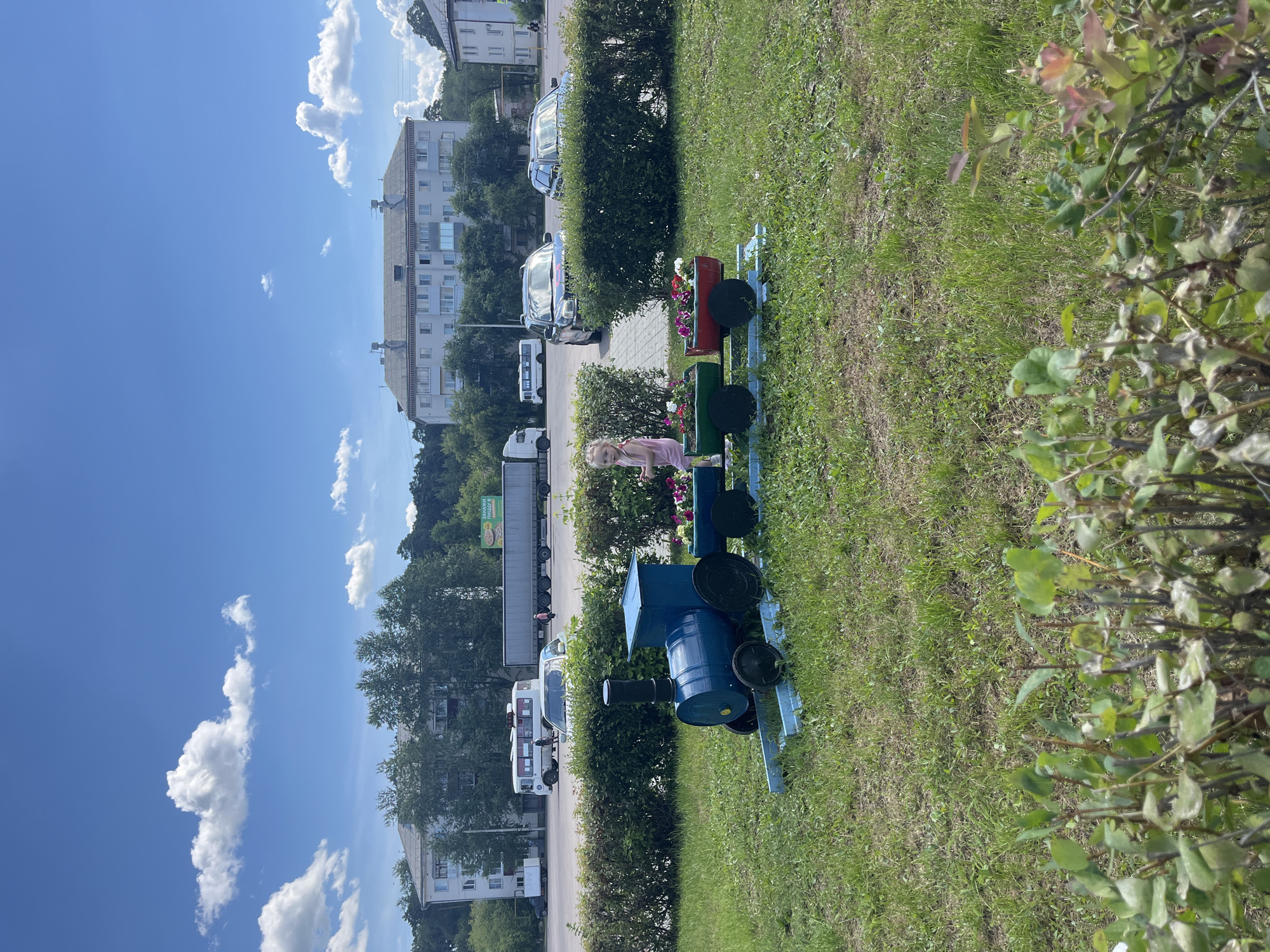 The image shows a miniature train flower planter decorated with flowers, situated in a grassy area next to a sidewalk and a road. A young girl in a pink dress is standing near the train, looking towards the camera. In the background, there are several buildings, including apartment blocks and a commercial building with a visible sign. Cars, trucks, and a bus are parked or driving on the road. The scene appears to be outdoors on a sunny day with a blue sky and scattered clouds. The presence of apartment buildings and a road suggests an urban or suburban setting, possibly in Shadrinsk, Russia, as indicated by the location context. The overall atmosphere is peaceful and sunny, with a touch of childlike playfulness due to the train planter and the girl's presence.