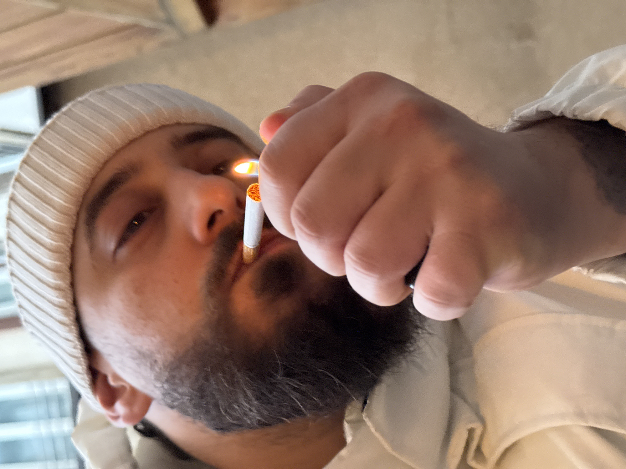 A close-up, angled photograph depicts an individual in the process of lighting a cigarette indoors. The person, viewed from a slightly low perspective, has dark eyes, a full dark beard, and a dark mustache. They wear a light-colored, possibly off-white or beige, ribbed knit beanie and a light-colored garment, potentially a jacket or hoodie. A white cigarette is held in the mouth, with its tip visibly ignited. The individual's right hand, prominently featured in the lower center, holds a black lighter that produces an active orange flame directed at the cigarette. The background is blurred and out of focus, showing indistinct light-colored surfaces and hints of what may be a window in the upper left. This scene is located in Strasbourg, France.