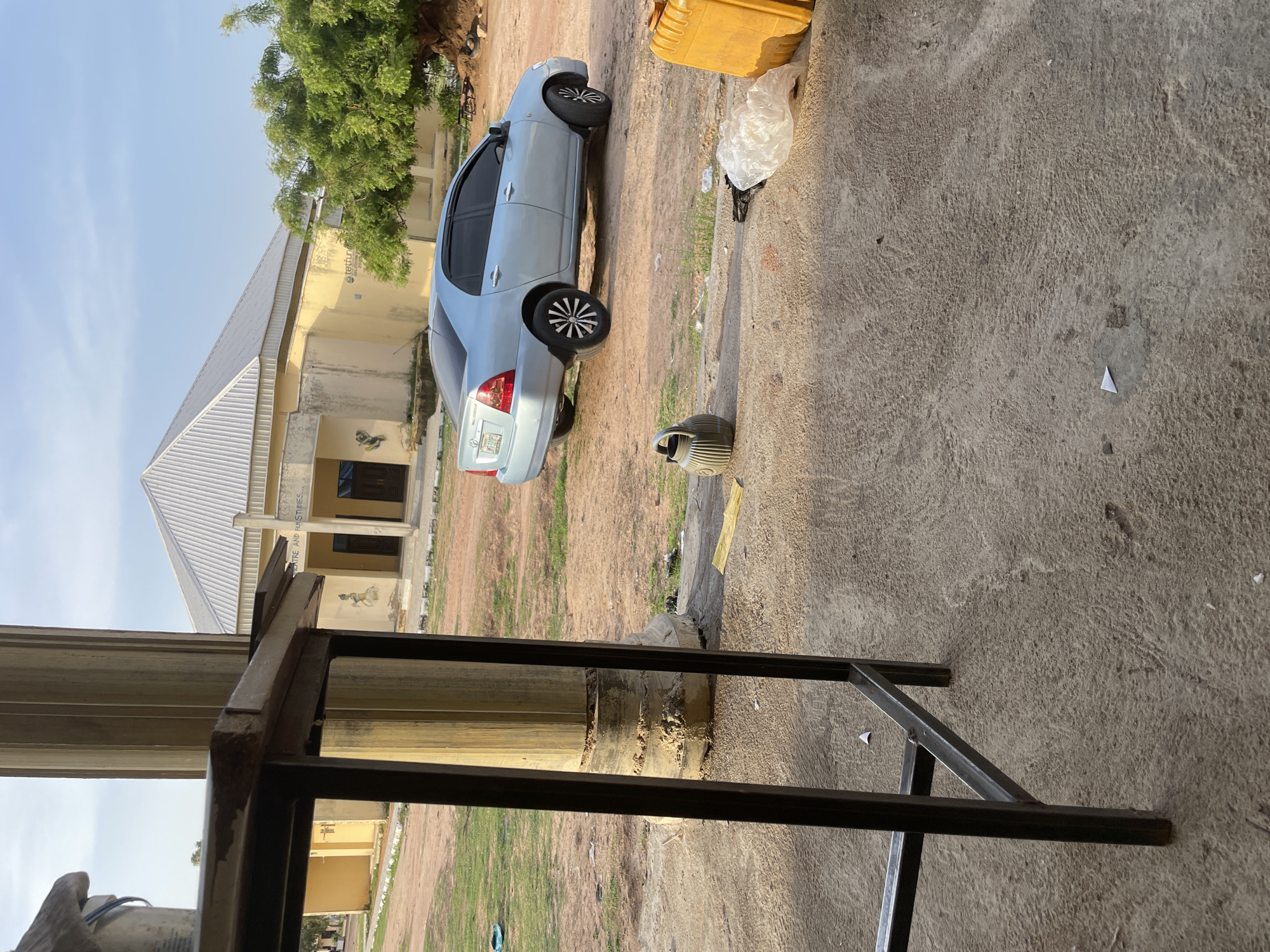 The image depicts an outdoor scene in Jalingo, Nigeria, featuring a light blue sedan parked on a dirt and concrete ground. The car is facing to the left of the frame. In the background, a yellow building with a silver, ribbed roof is visible, and a large tree with green foliage stands to the right of the building. A black metal table frame is positioned in the foreground, obscuring part of the view. Scattered on the ground are various objects including a gray watering can, a plastic bag, and a yellow container. The sky above is a clear blue with no visible clouds. The lighting suggests it is daytime, likely late afternoon given the shadows.