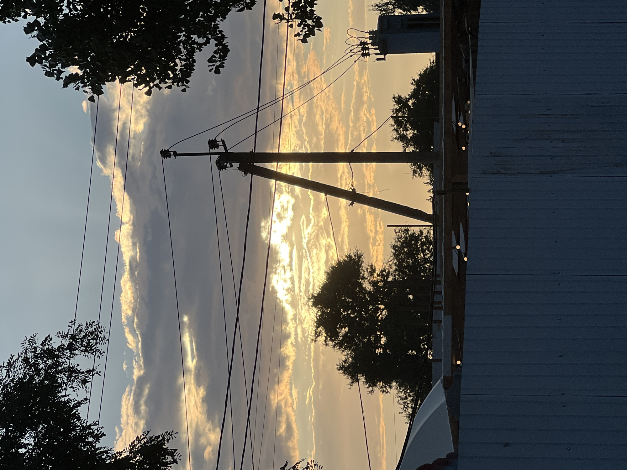 A silhouetted view of the sky over Golubitskaya, Russia, at sunset. The sky is filled with clouds, some of which are illuminated by the setting sun, creating streaks of light. Power lines crisscross the frame, and a utility pole stands prominently in the center. In the foreground, a white fence is visible, and beyond it, the silhouettes of trees and structures. The overall scene is one of a rural or suburban setting at dusk, with elements of human infrastructure against the backdrop of a natural phenomenon.