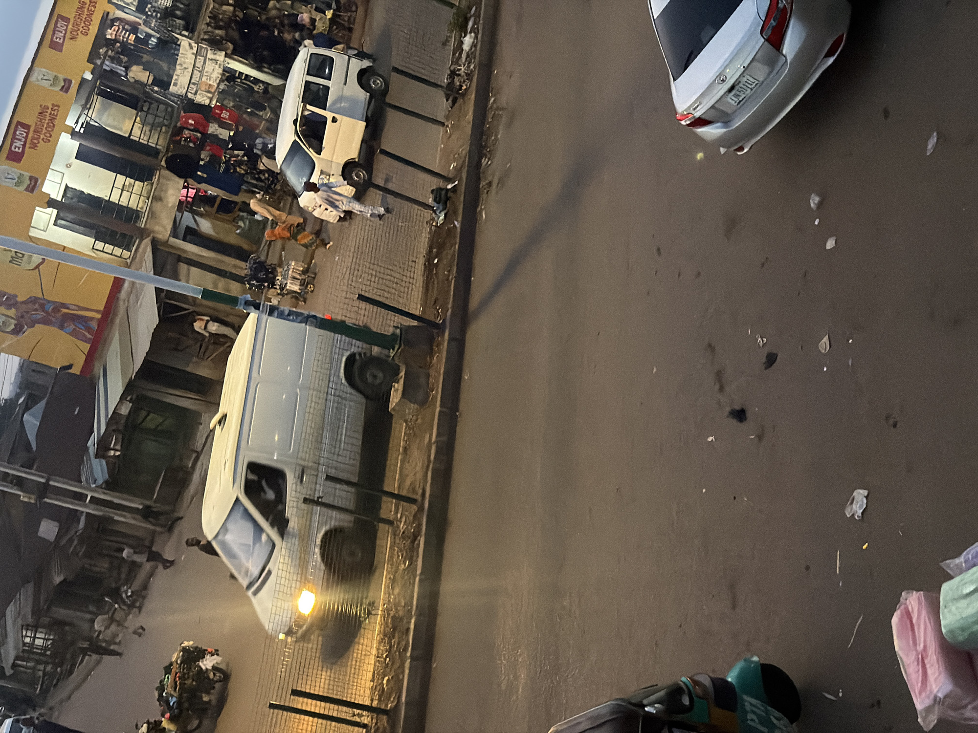 This is a ground-level, slightly angled shot of a street scene in Bukuru, Nigeria, likely at dusk or night, as indicated by the streetlights and the overall dim lighting. The scene depicts a bustling market or commercial area with several vehicles, including a white van and a silver car. A pile of trash is visible on the left side of the frame.
In the foreground on the left, a white van is parked with its door open, and a person appears to be interacting with it. Behind the van, a storefront with yellow and brown signage is visible. The signage includes text such as "ENJOY NOURISHING GOODNESS" and what appears to be a partial brand name or slogan. More stalls and shops with various goods are visible in the background, suggesting a lively marketplace.
To the right, a silver car is parked on the street. Its license plate, though partially obscured and blurry, seems to read "LM51LTL". The street is paved, and the area appears to be an urban environment. The presence of people, vehicles, and merchandise points to ongoing commercial activity. The trash pile suggests waste management challenges or a busy, perhaps less regulated, area. The overall impression is one of a vibrant, everyday scene in a Nigerian town.