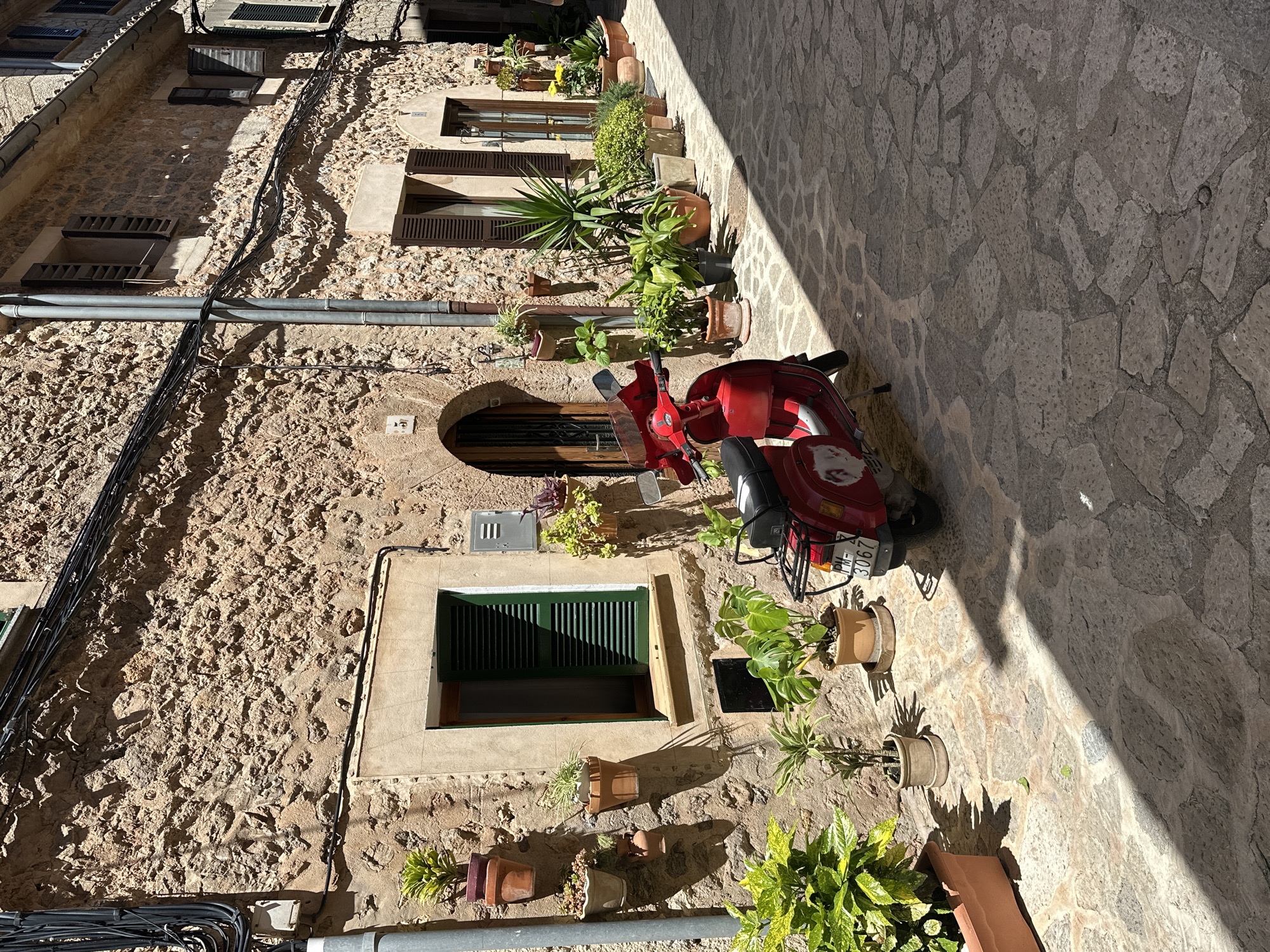 The image captures a narrow cobblestone street in Valldemossa, Spain. A red Vespa scooter is parked along the wall, which is constructed of rough-hewn stone. Several potted plants line the wall, some placed directly on the ground, others hanging near windows. The windows are varied, some with dark wooden shutters, others with green, and one shaped as an arch. Electrical cables run along the upper part of the wall. The street is partially shaded.