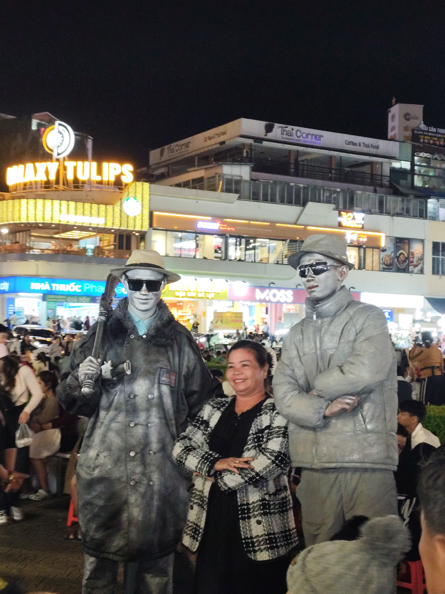 The image depicts a nighttime scene in Da Lat, Vietnam. Two street performers with silver body paint, wearing hats and sunglasses, stand alongside a woman in a black dress and a black and white plaid jacket. The performer on the left is holding a tool resembling a pickaxe. They are likely posing for a photo with the woman. The background features brightly lit buildings, including establishments such as "Galaxy Tulips," "Thai Corner," "Moss," and "Nhà Thuốc Pha." People are visible in the background, suggesting a crowded outdoor setting.