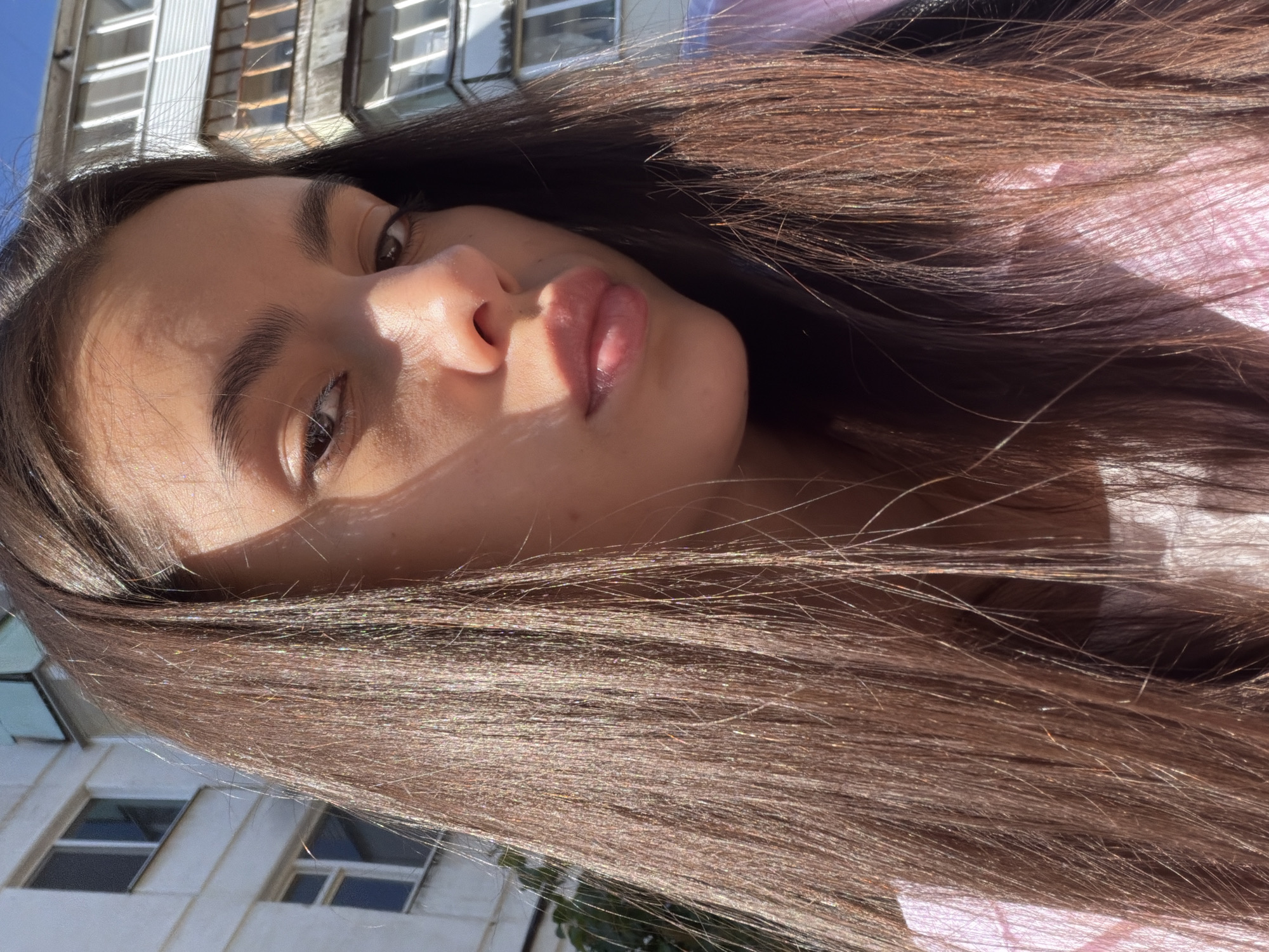 A close-up selfie captures a young woman with long, dark brown hair and brown eyes. She is looking towards the camera with a neutral expression. The background consists of a residential building with balconies and windows, suggesting an urban environment. The lighting indicates it is daytime, with sunlight casting shadows across her face. The building appears to be a multi-story apartment complex, typical of Soviet-era architecture, possibly located in the city of Chaikovsky, Russia, given the context.