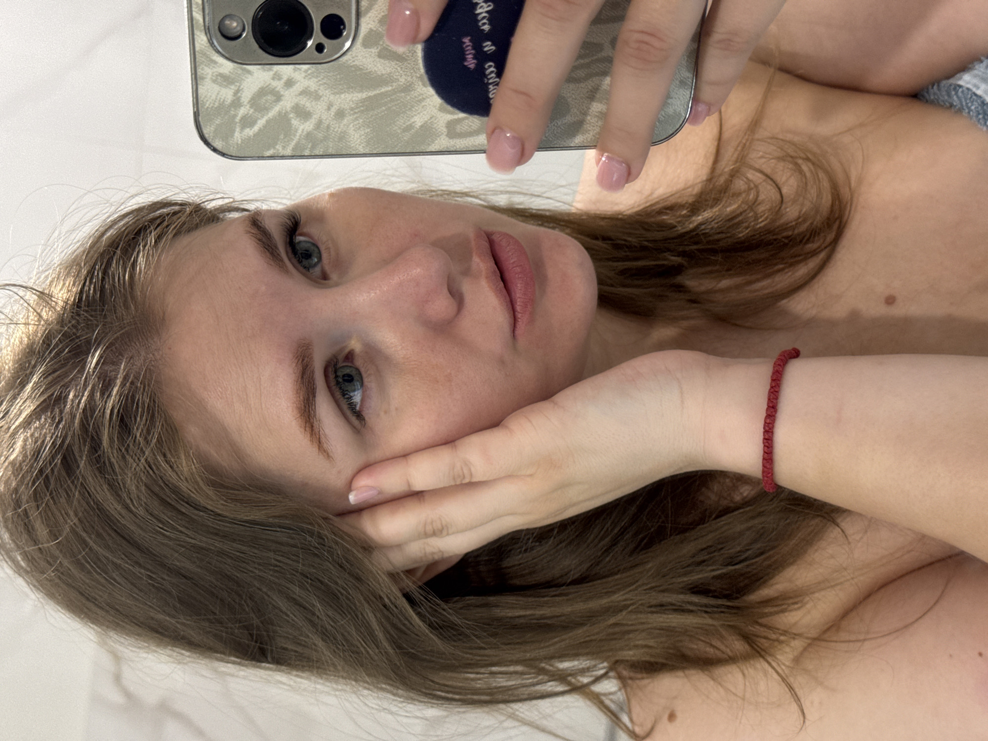 A woman is taking a selfie indoors. She holds a smartphone with a light-colored patterned case in her right hand. Her left hand is resting against her cheek. She has light brown hair and light blue eyes. A red bracelet is on her left wrist. Her fingernails are painted with a pale pink polish. Part of her shoulder and chest are visible. The background appears to be a white, tiled wall. There is no evidence that the image was taken in the city of Ферма, Russia.