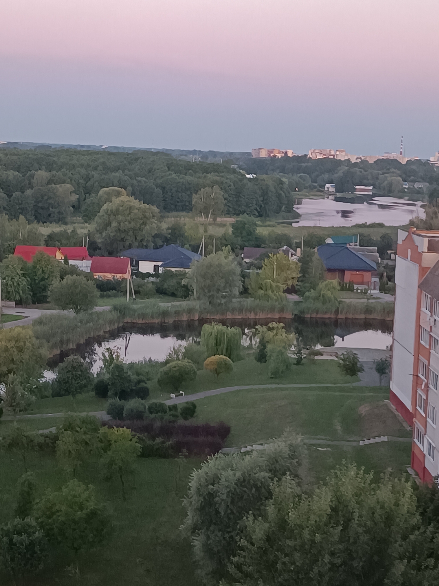 This is an aerial, wide-angle shot of a residential area on the outskirts of Brest, Belarus, during dusk. The sky exhibits a gradient of soft pink and light blue, indicative of twilight. In the distance, a cluster of mid-rise apartment buildings with visible windows and balconies are silhouetted against the horizon. Closer to the foreground, a dense, green forest borders a tranquil body of water, likely a river or lake, which reflects the muted sky. Several modest houses with pitched roofs, some in red and others in dark blue and white, are nestled among the trees and greenery. A paved path winds through a park-like setting dotted with various trees and bushes. The edge of a modern apartment building, featuring a light-colored facade with red accents and evenly spaced windows, is visible in the lower right quadrant of the image. The scene is serene and appears to be a typical residential landscape.