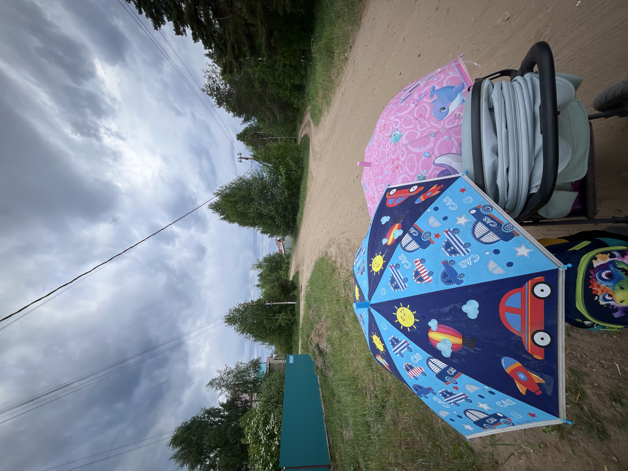 The image shows a dirt road in Chaikovsky, Russia, with a stroller containing a folded carriage and two children's umbrellas. One umbrella is pink with a whale design, and the other is blue with cars, rockets, and suns. A backpack with a colorful design sits next to the stroller. Overcast skies are visible above trees along the road. Power lines are also visible. A section of a teal fence is in the foreground.