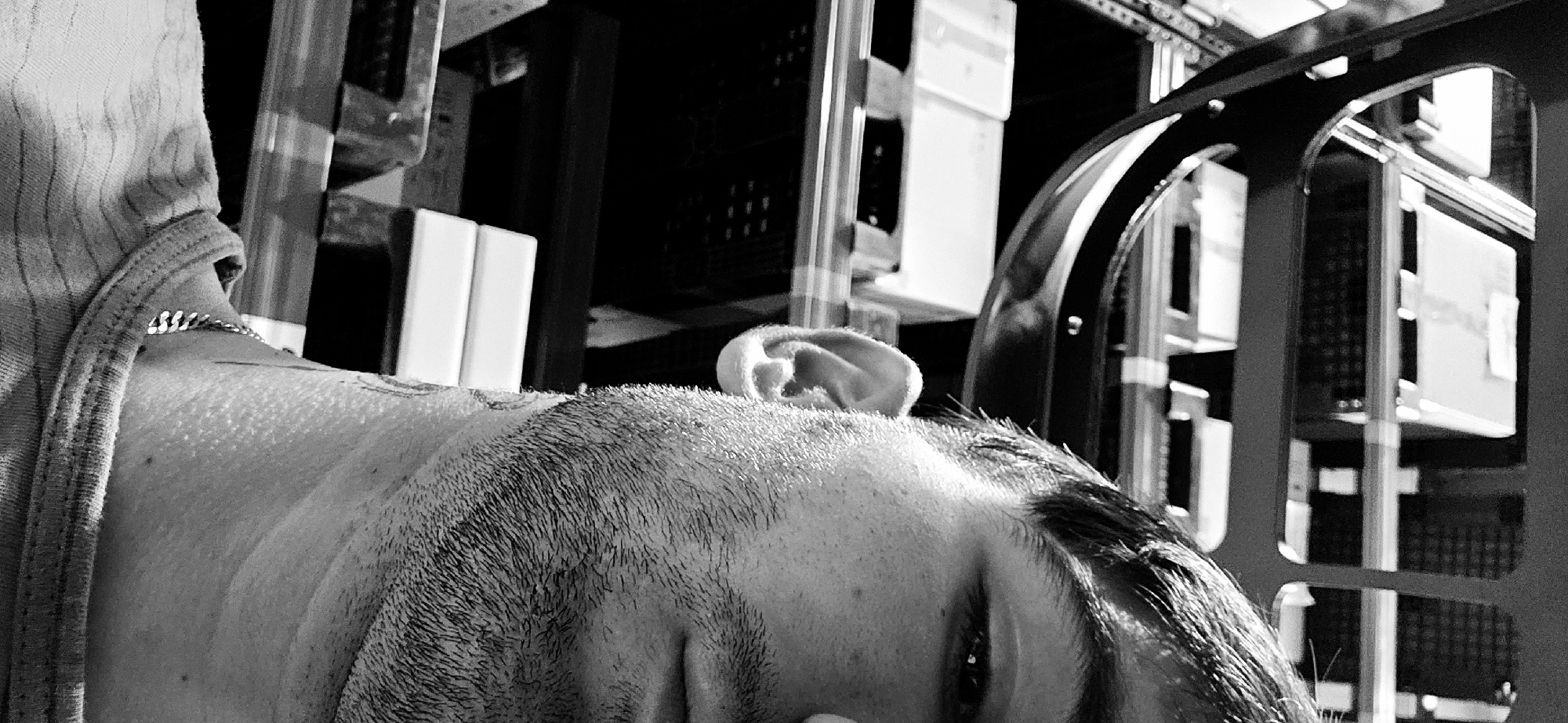 A black and white close-up image shows a man lying down. The man's head and upper chest are visible. He has short hair and a beard. Part of his face, including an eye and his ear, are visible. He is wearing a shirt with a striped pattern. He has a chain around his neck and a tattoo on his upper chest. The background includes various pieces of equipment, some of which appear to be part of a rack or shelving unit. The image is taken from a low angle. The location in Plovdiv, Bulgaria cannot be determined based on the image alone.