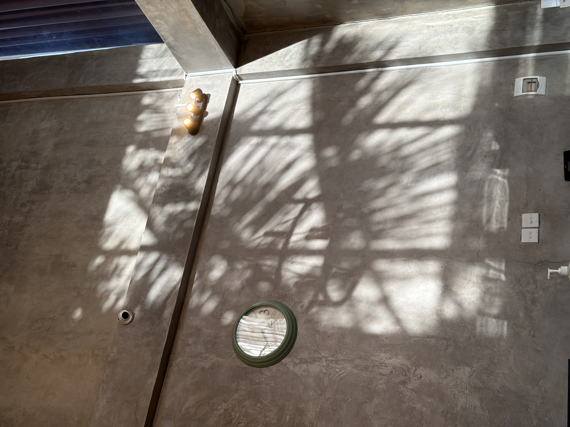A high-angle shot depicts an interior wall in North Kuta, Indonesia, bathed in sunlight and shadow. The textured grey concrete wall is bisected by a supporting beam. A round, green-framed clock is mounted on the lower section of the wall. A light fixture with three lights is affixed to the wall near the beam. Shadows from foliage create patterns on the wall. Electrical outlets and a light switch are installed on the right side of the wall. A small circular security camera is on the opposite side of the wall, to the left of the clock. An emergency exit sign is near the upper right of the wall. A white linear fixture runs along the top edge of the wall.
